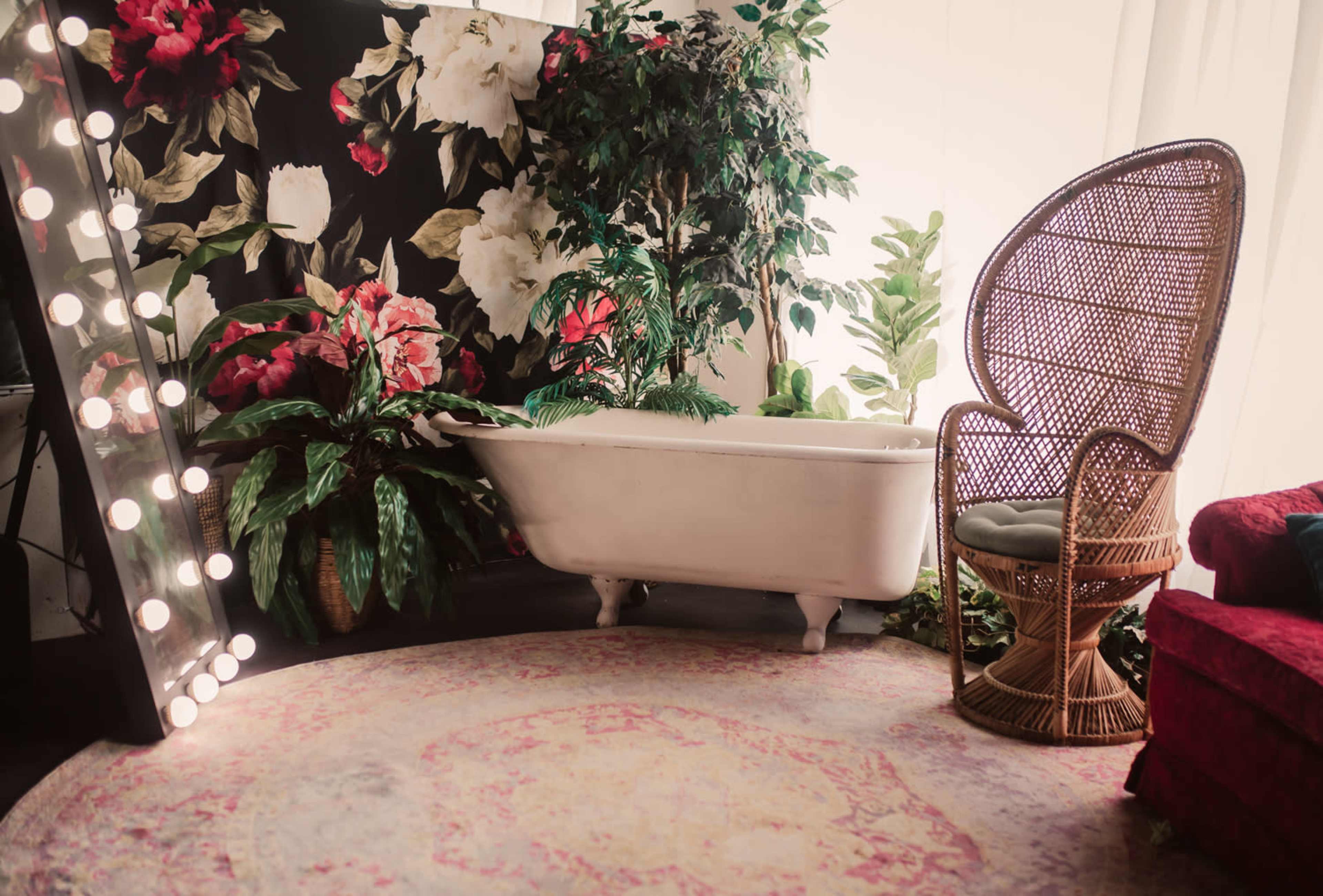 A vintage white bathtub alongside a lush backdrop of flowers, a rattan chair, and decorative plants in a cozy interior space.