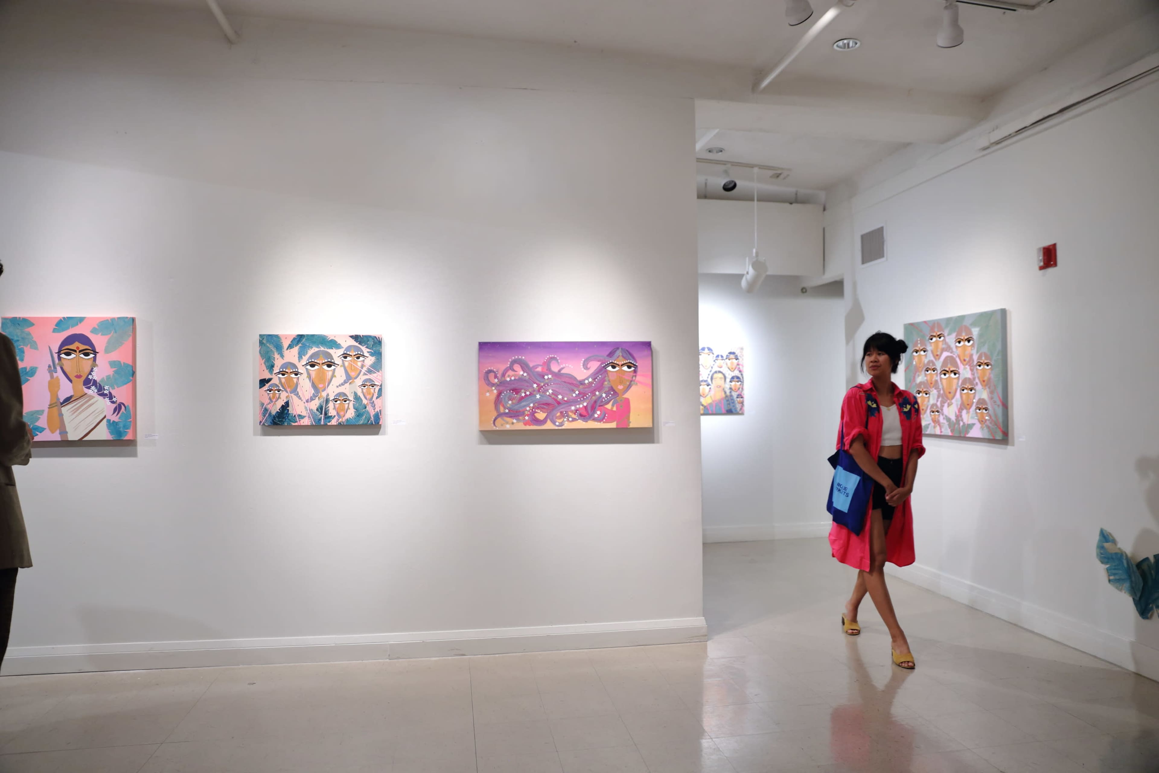 A woman in a pink dress walks past colorful paintings on the walls of an art gallery.
