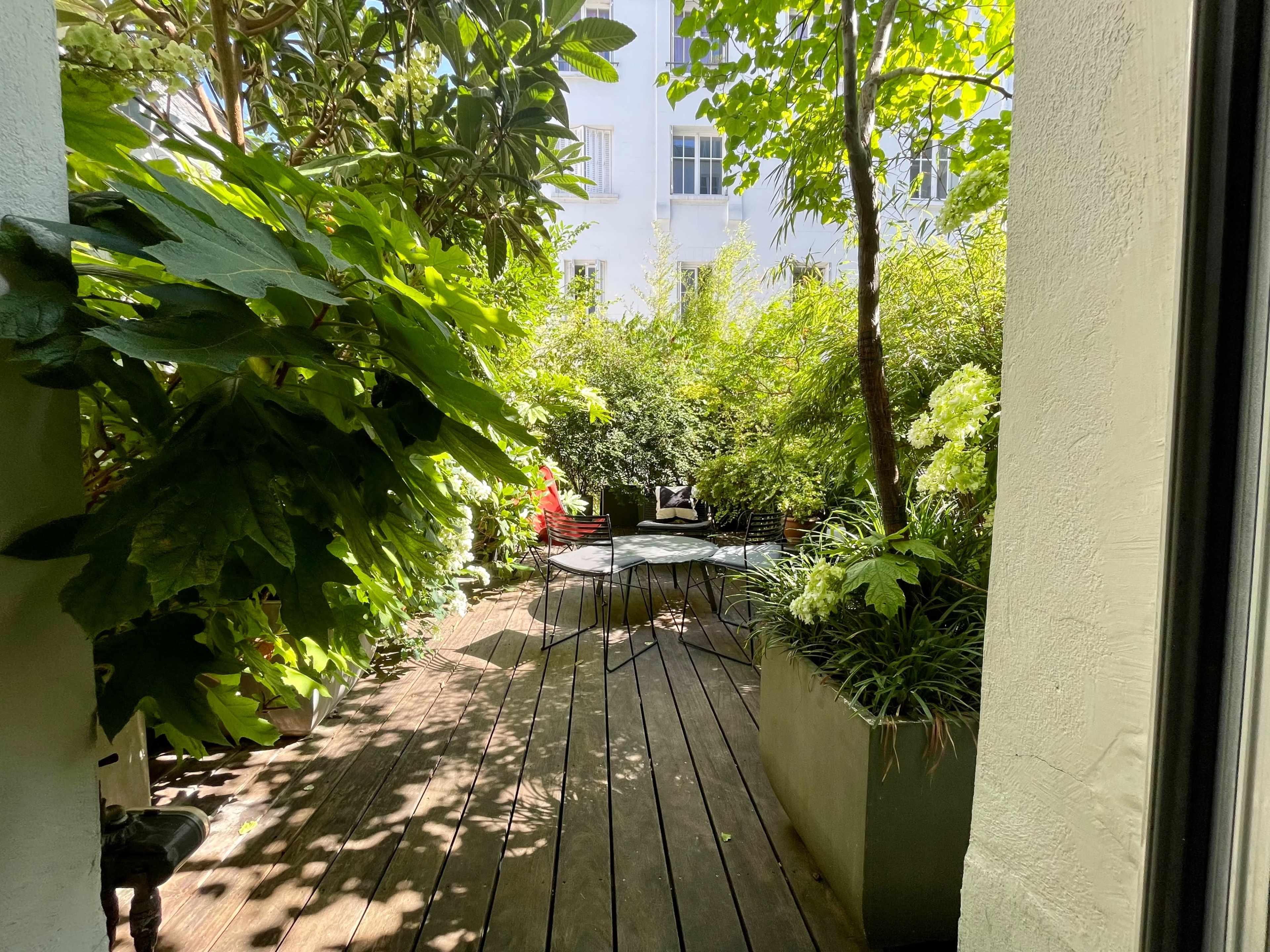 A wooden deck surrounded by lush greenery leads to a small seating area in a garden.