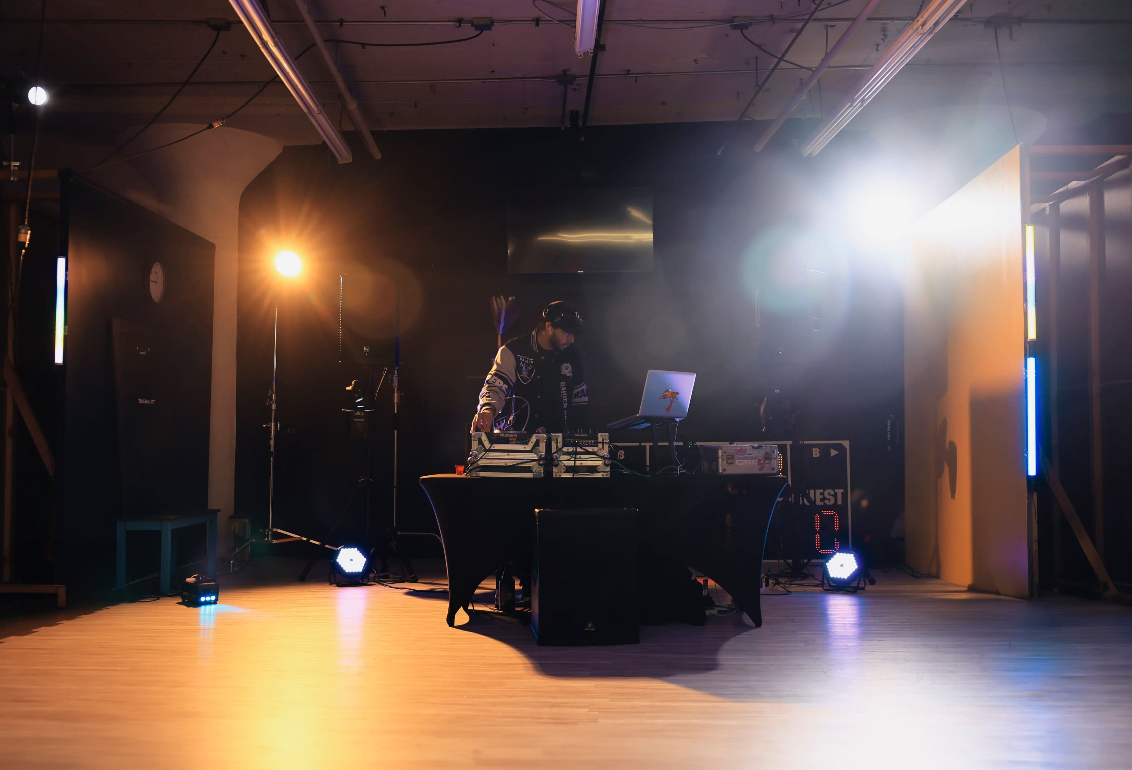 A DJ stands behind a table with turntables and a laptop, illuminated by stage lights in a dimly lit venue.