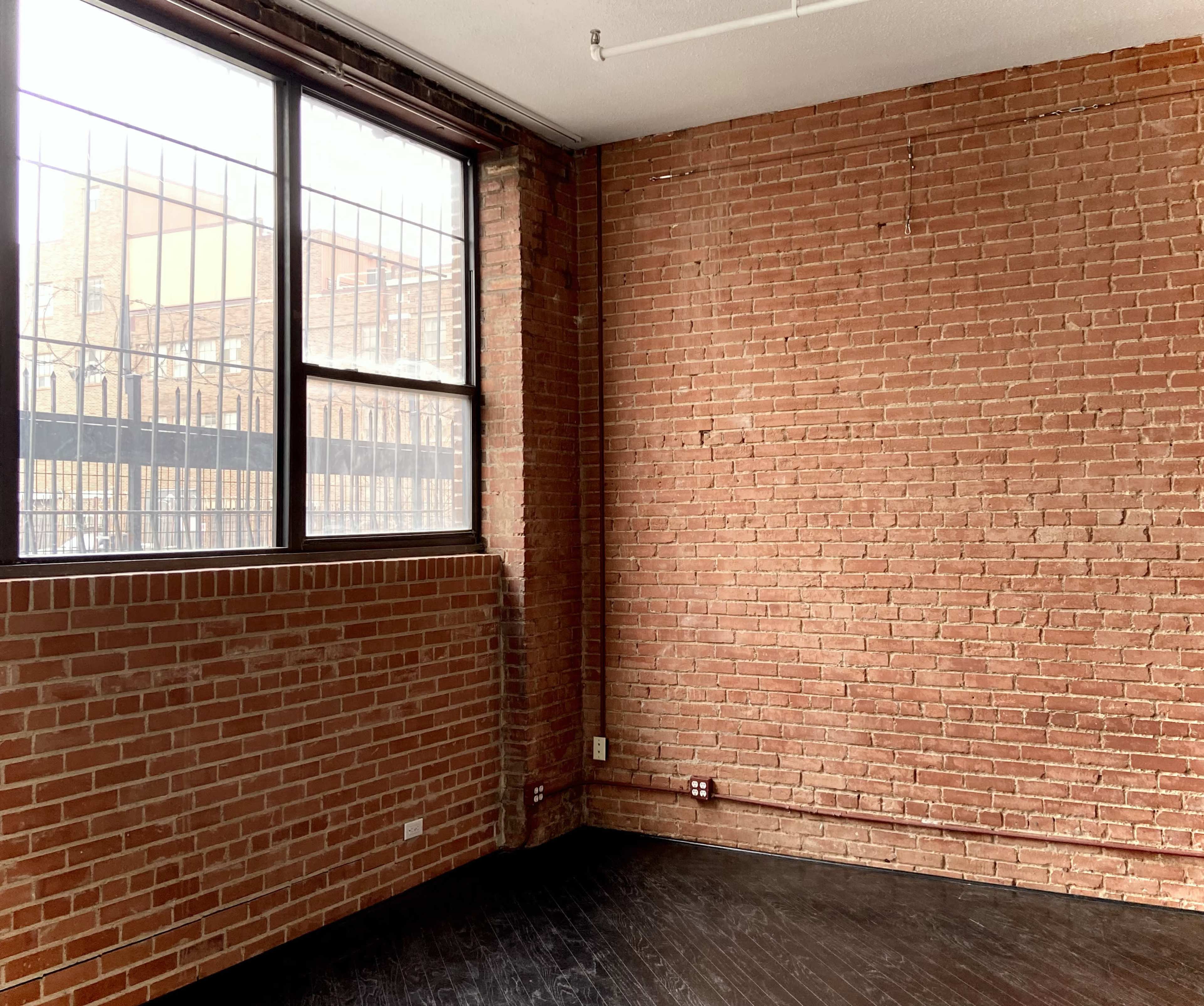 The image shows a vacant room with exposed brick walls, a large window, and a dark wooden floor.