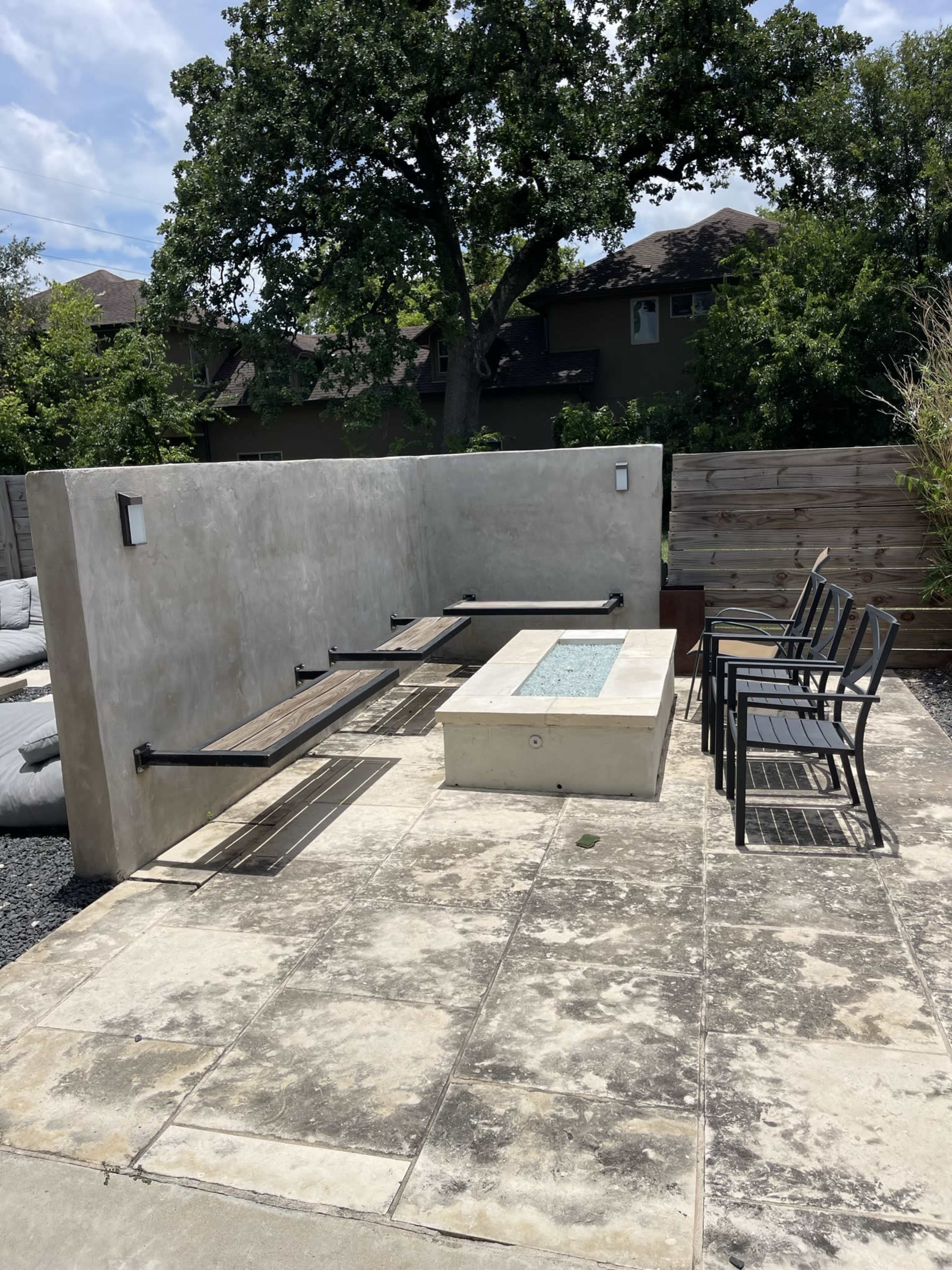 A concrete seating area with a narrow fire pit is accompanied by several metal chairs against a backdrop of greenery and wooden fencing.