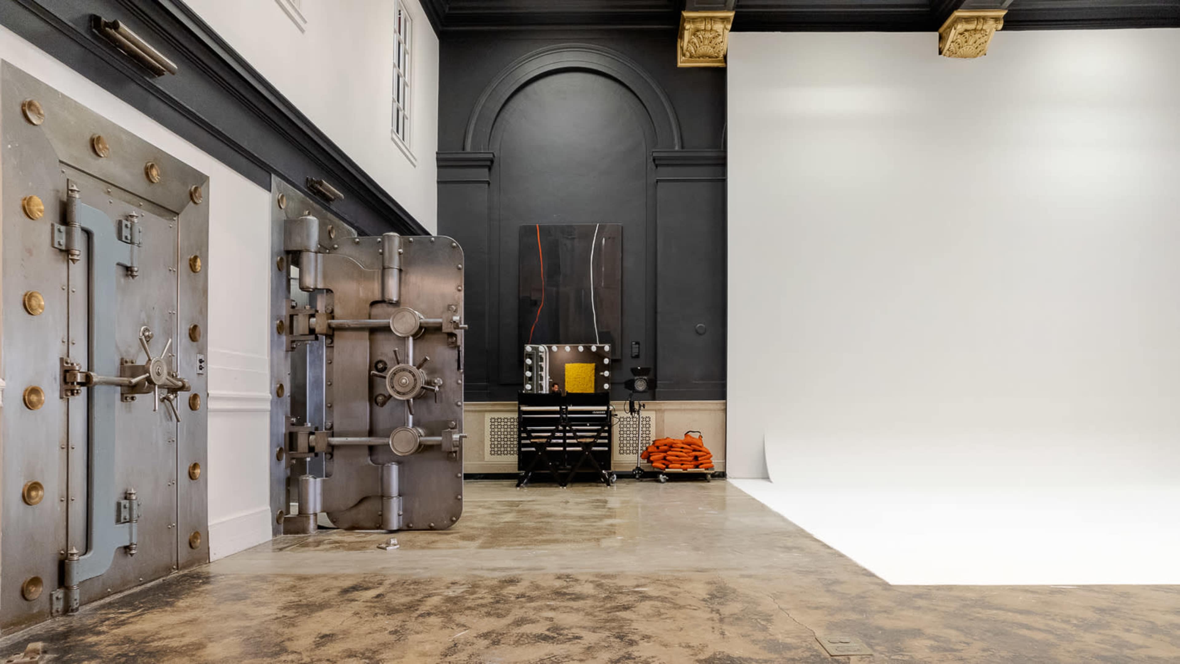 A spacious studio with a large, metallic vault door, a black wall with a painting, and a clean white backdrop on one side.
