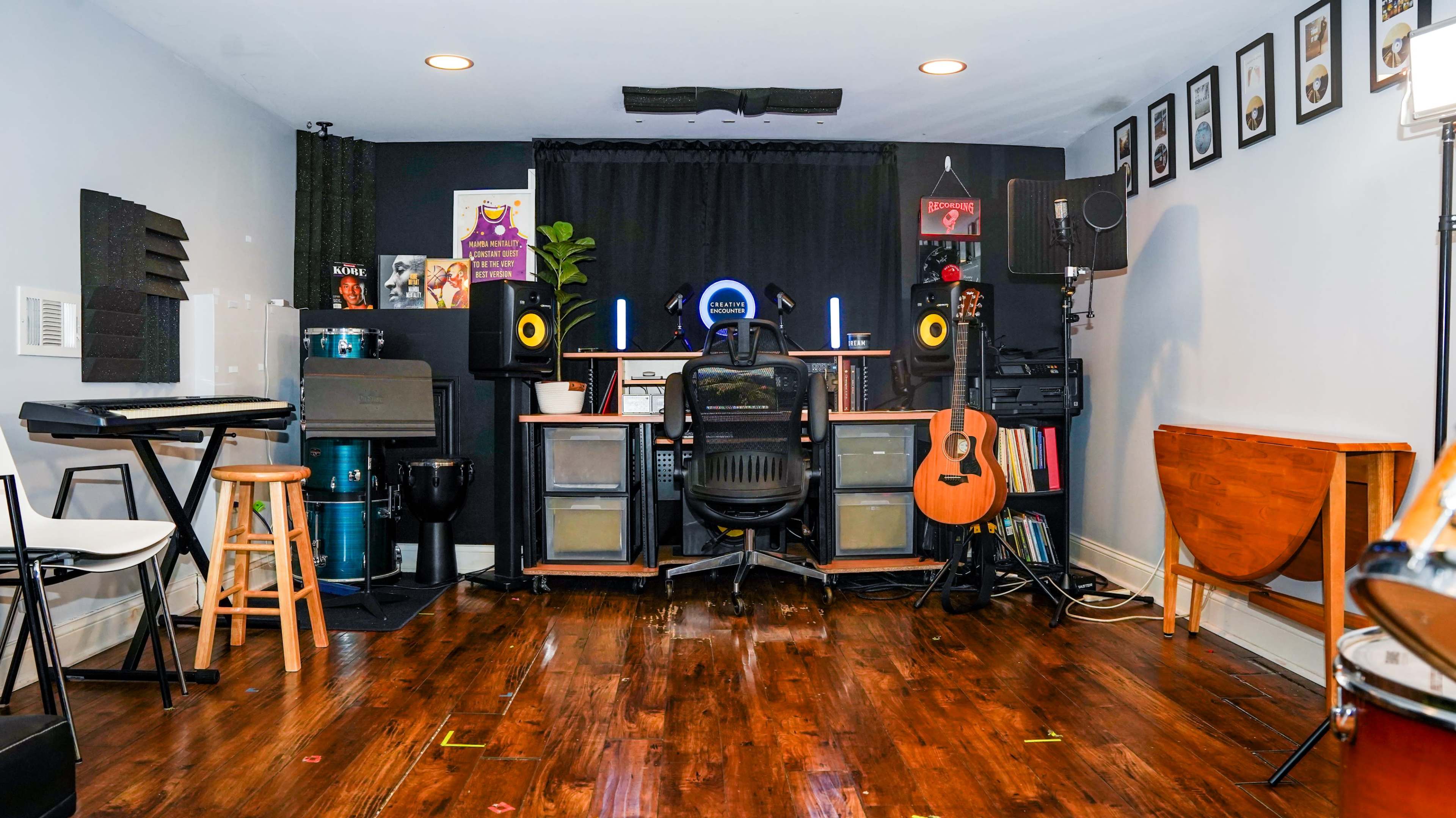 A music studio featuring a desk with computer equipment, speakers, a guitar, and various musical instruments arranged around the room.