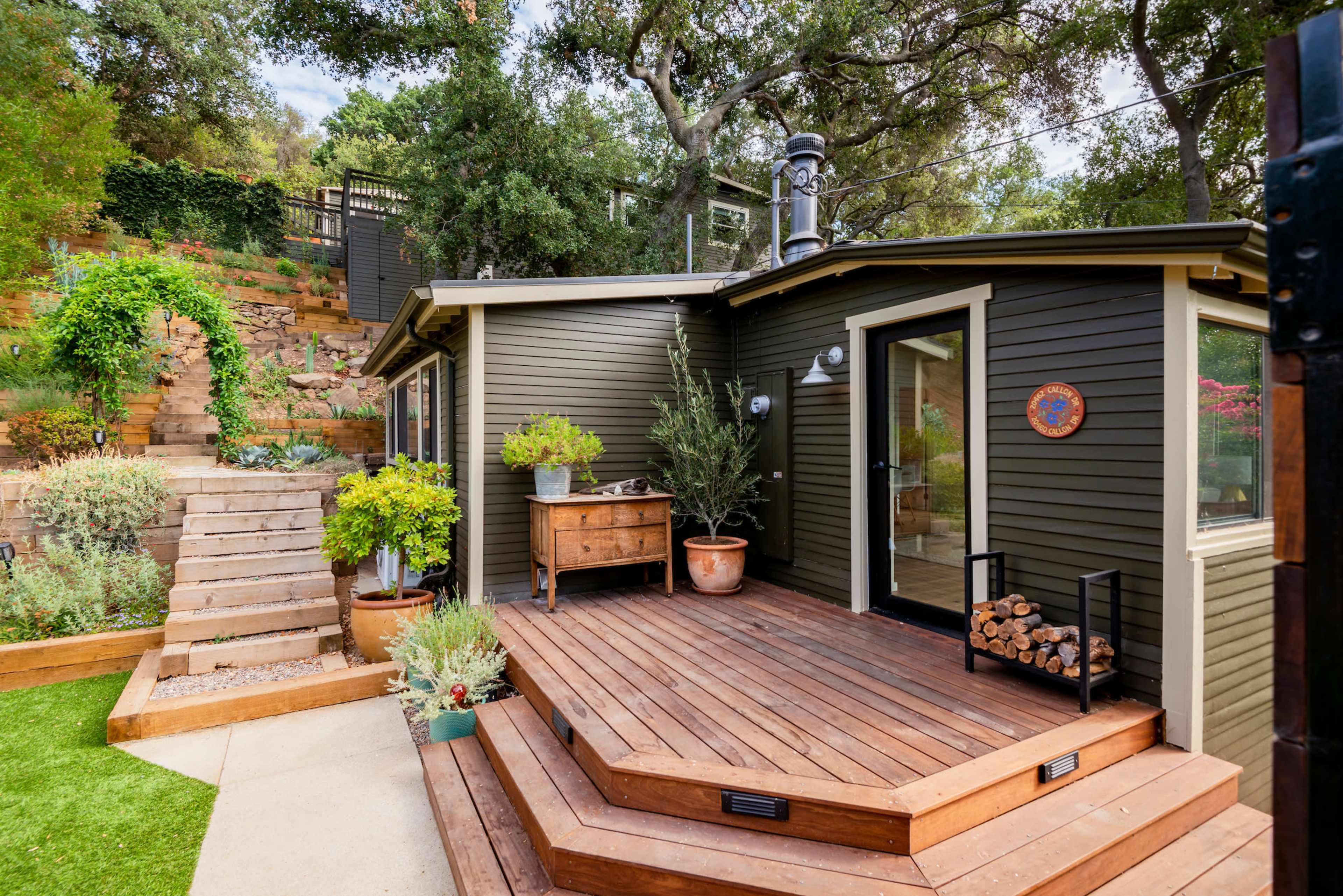 A small, modern wooden deck attached to a dark green house features a potted plant and a wood storage area, with stone steps leading up to a landscaped garden.