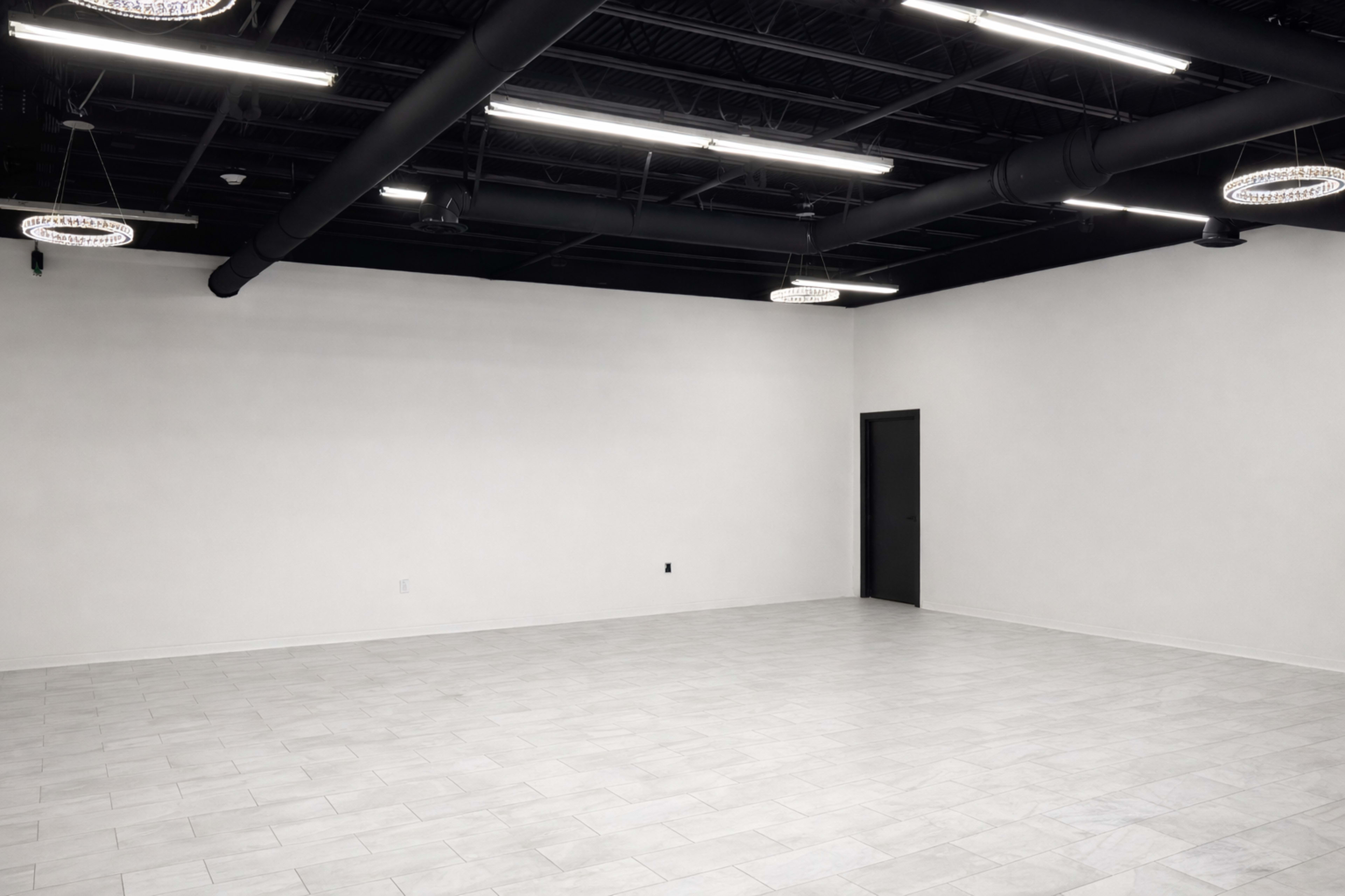 The image shows a spacious, empty room with white walls, tiled flooring, and black ceiling pipes with circular light fixtures.