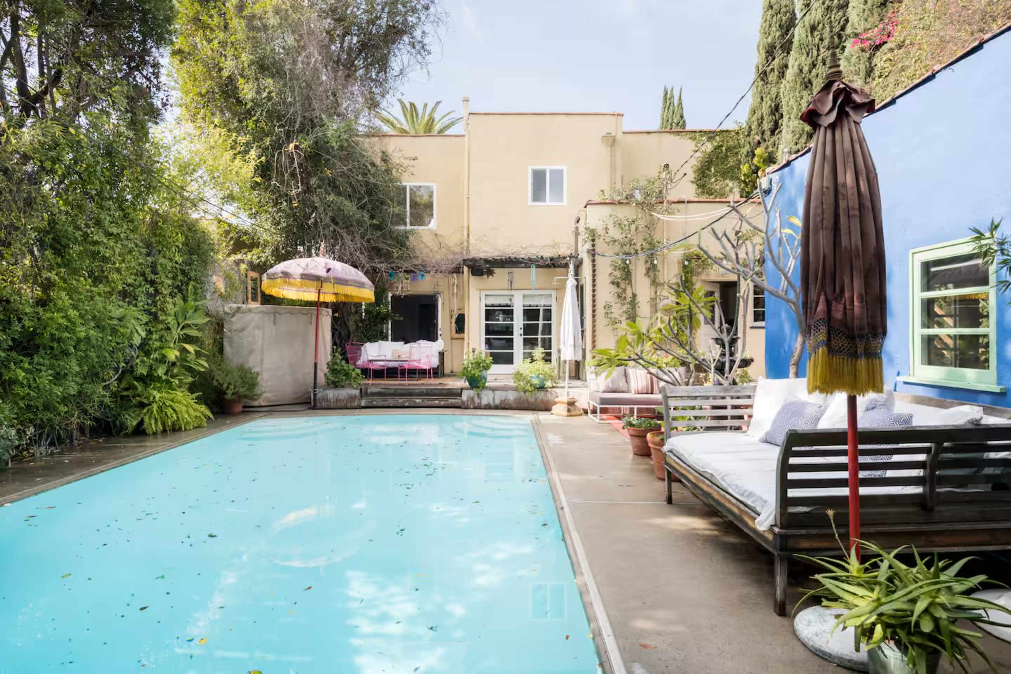 The image depicts a tranquil backyard scene featuring a swimming pool surrounded by lush greenery, with a blue wall and a patio area that includes outdoor seating and a colorful umbrella.
