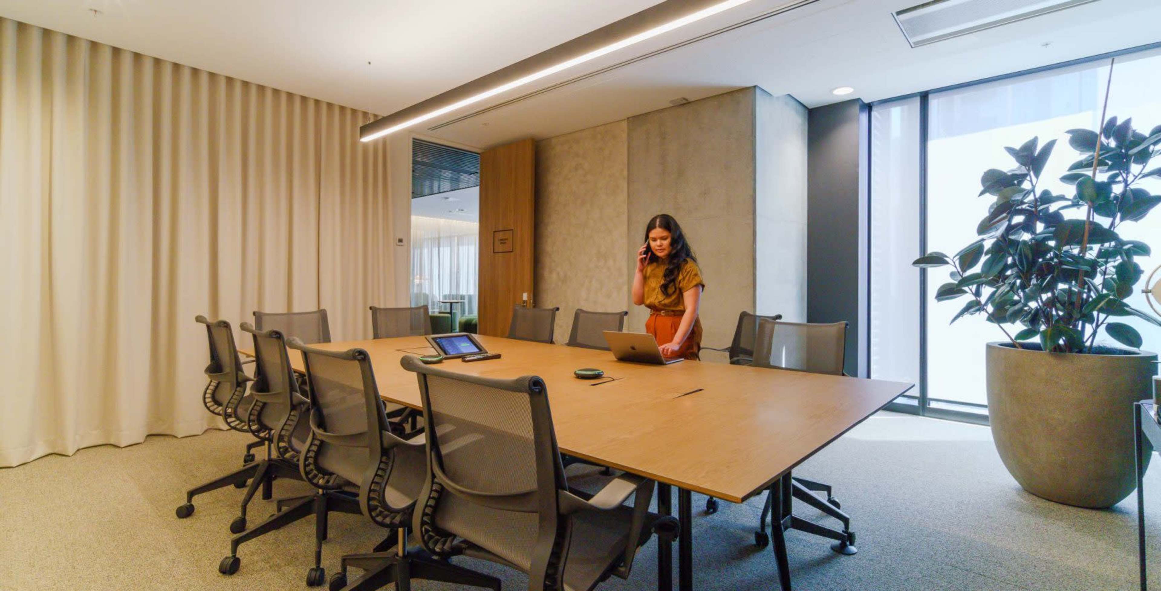 A woman stands in a conference room with a large wooden table surrounded by chairs, working on a laptop while holding a phone to her ear.