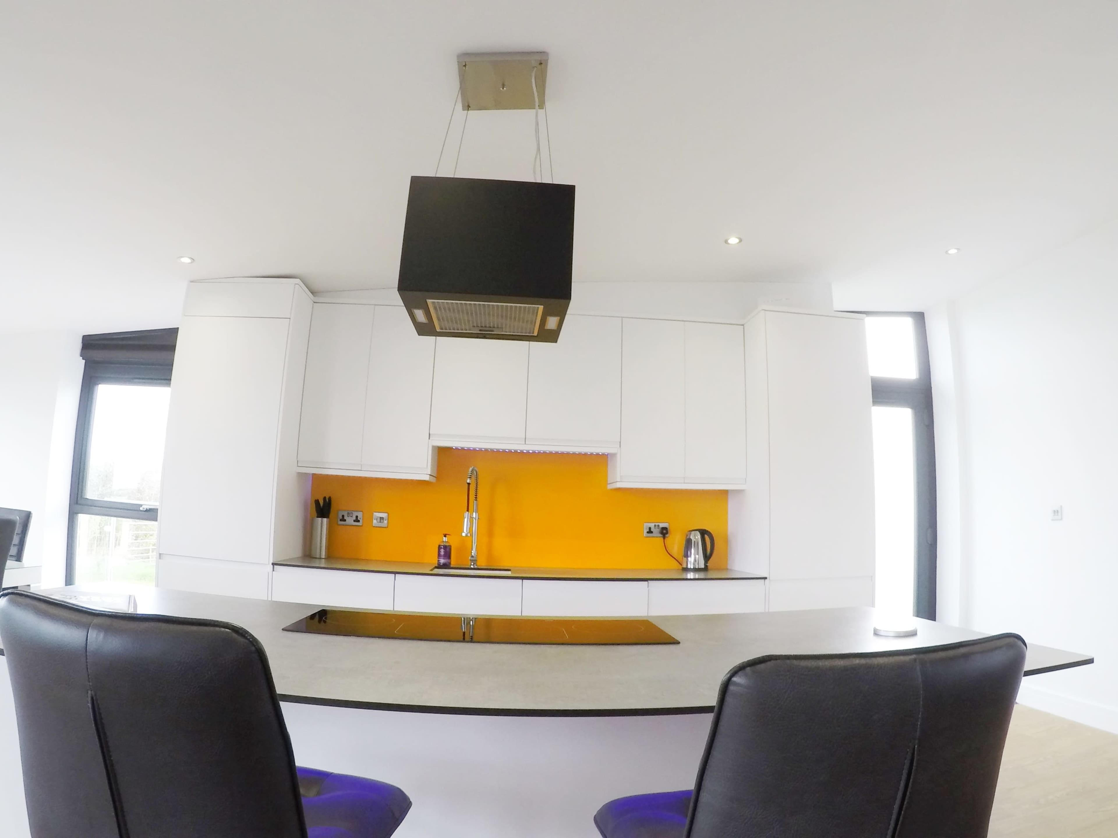 A modern kitchen features white cabinetry, a gray countertop, an orange backsplash, and a suspended black range hood overhead.