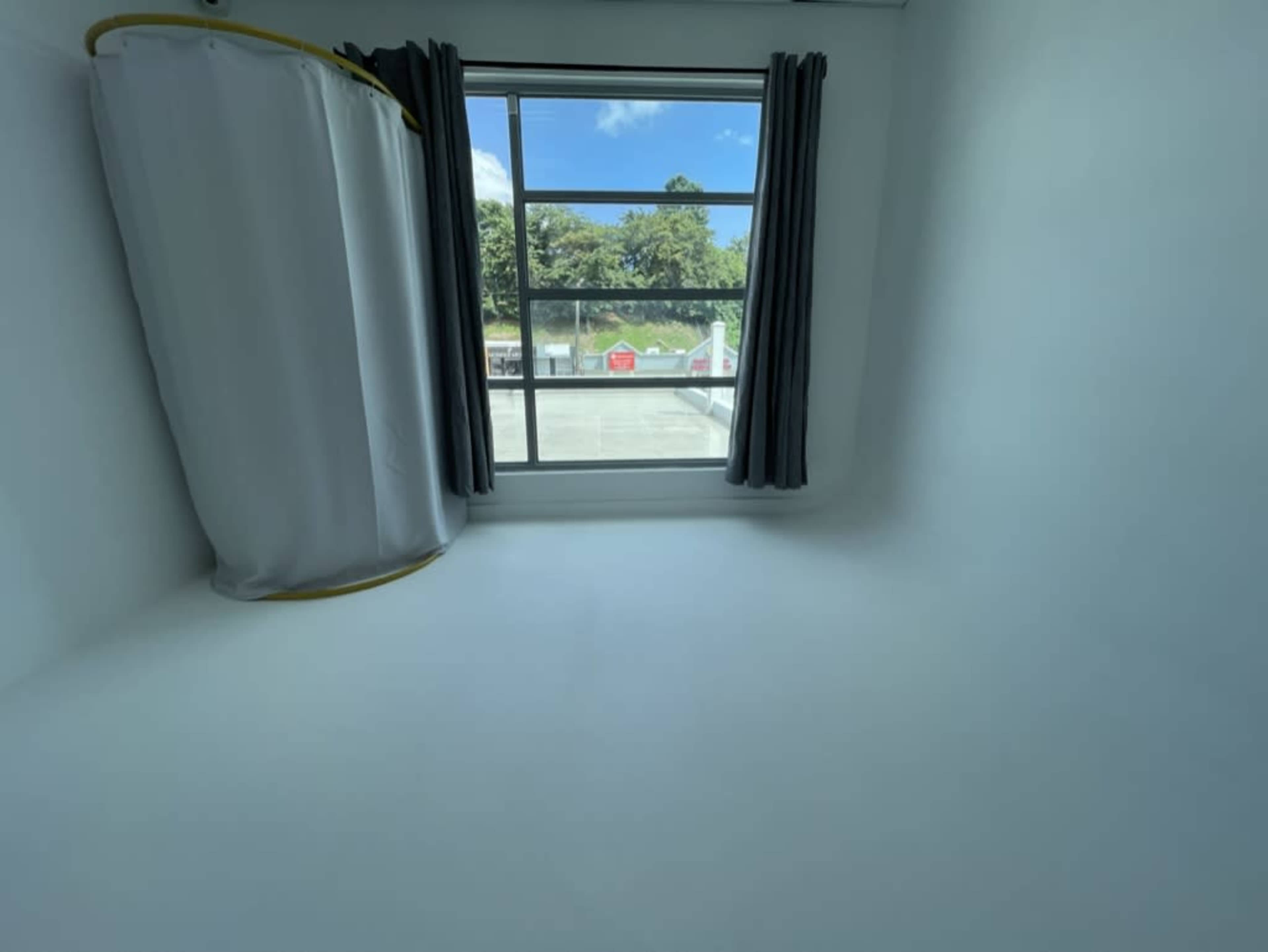 The image shows a sparse room with a window revealing a view of trees and a parking area outside, and a curtain rod hanging from the ceiling.