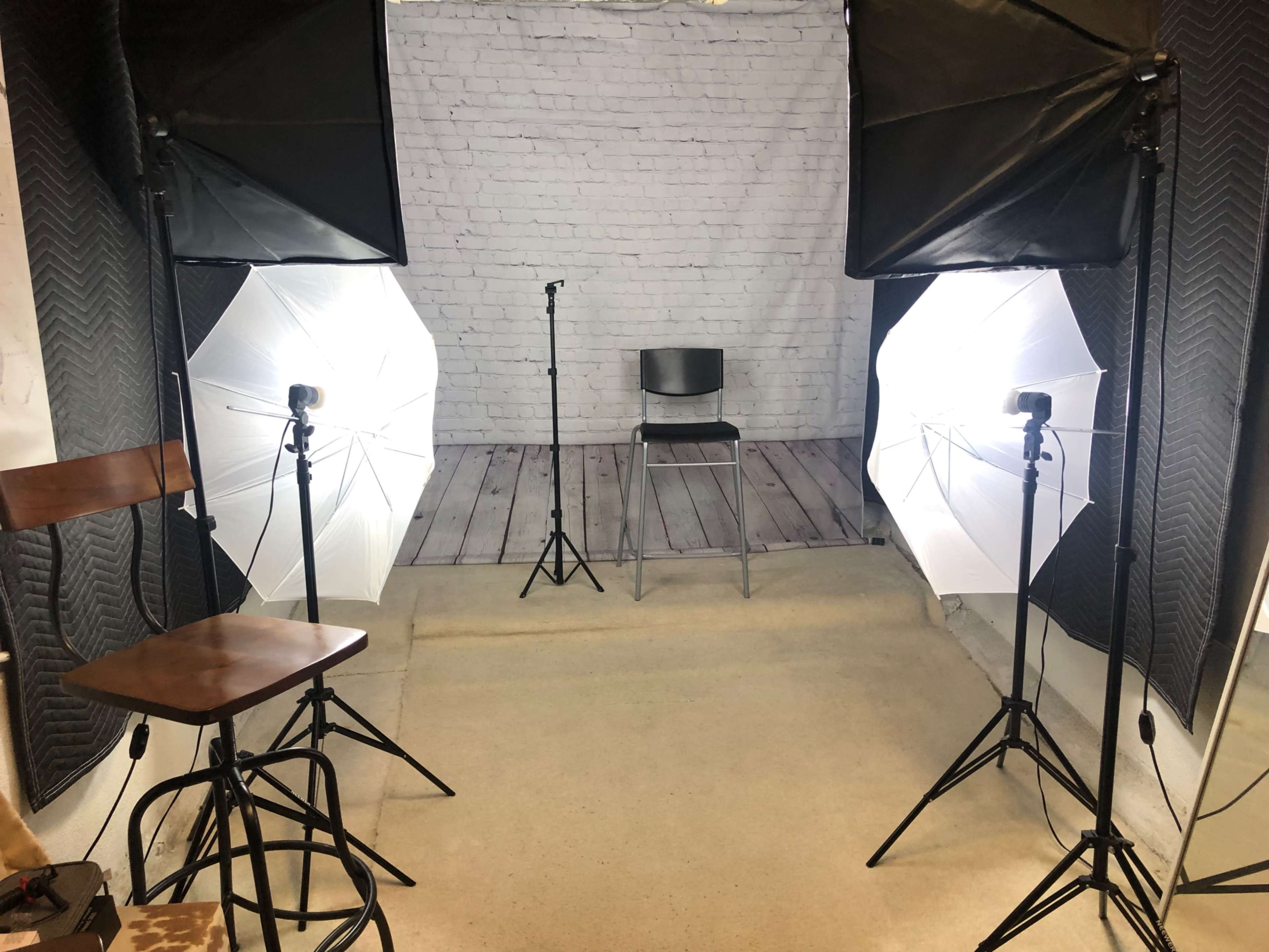A photo studio setup with two light stands, a black chair in the center, and a backdrop of white brick walls.