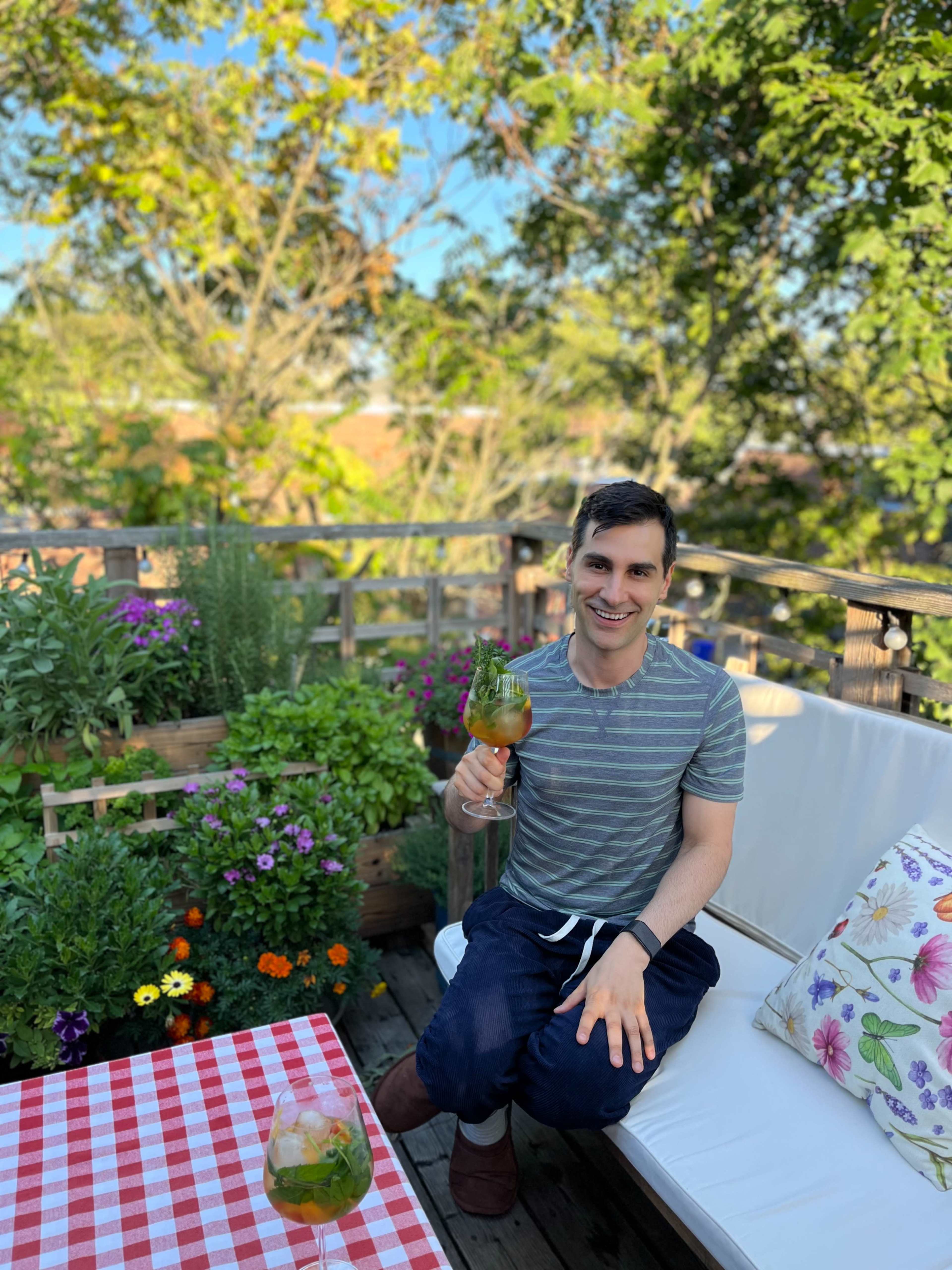 A man sits on a patio surrounded by colorful flowers, holding a drink, with a checkered tablecloth in the foreground.