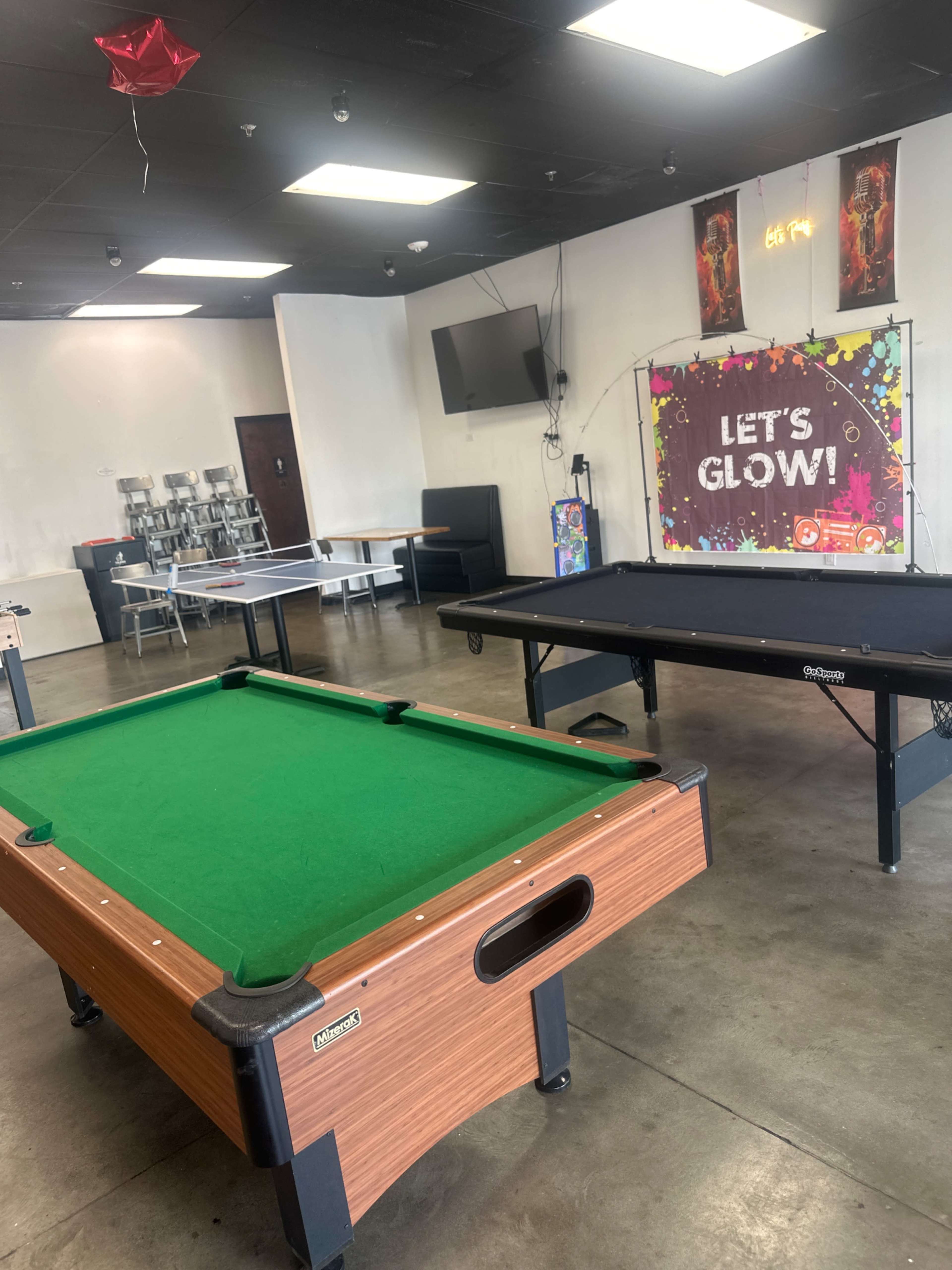 The image shows a recreational room with a green felt pool table on the left and a black ping pong table on the right, along with tables and a television in the background.