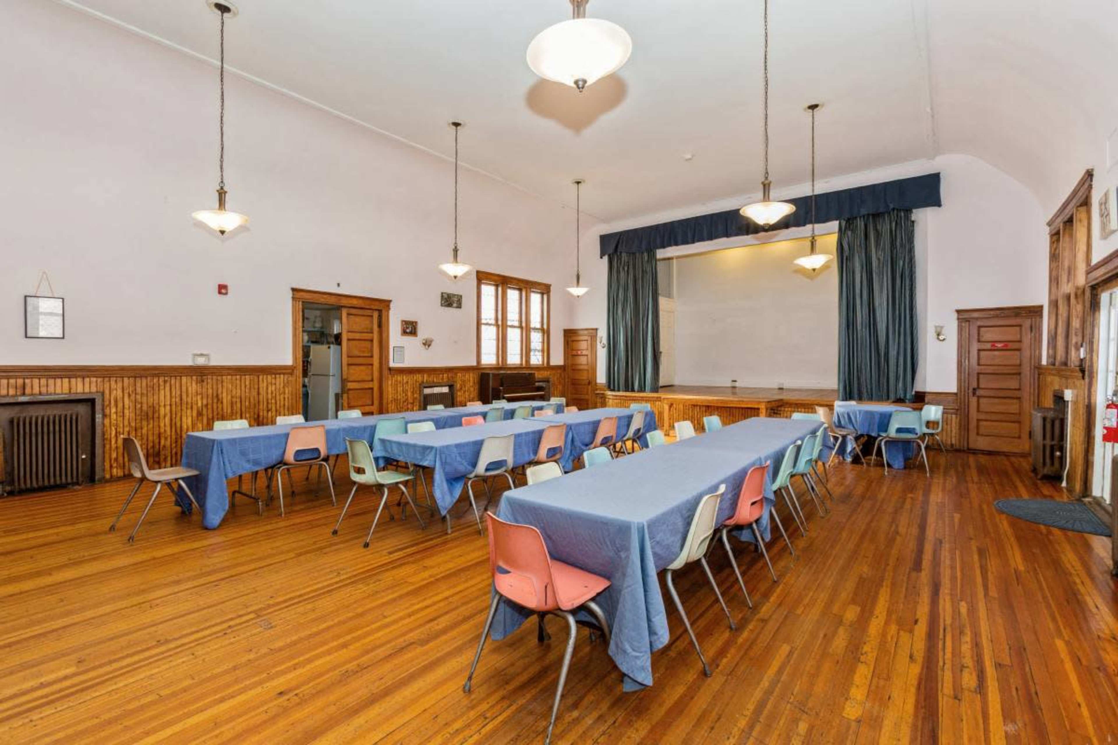 A spacious classroom with wooden floors, long tables covered in blue tablecloths, and chairs in various colors arranged around them.