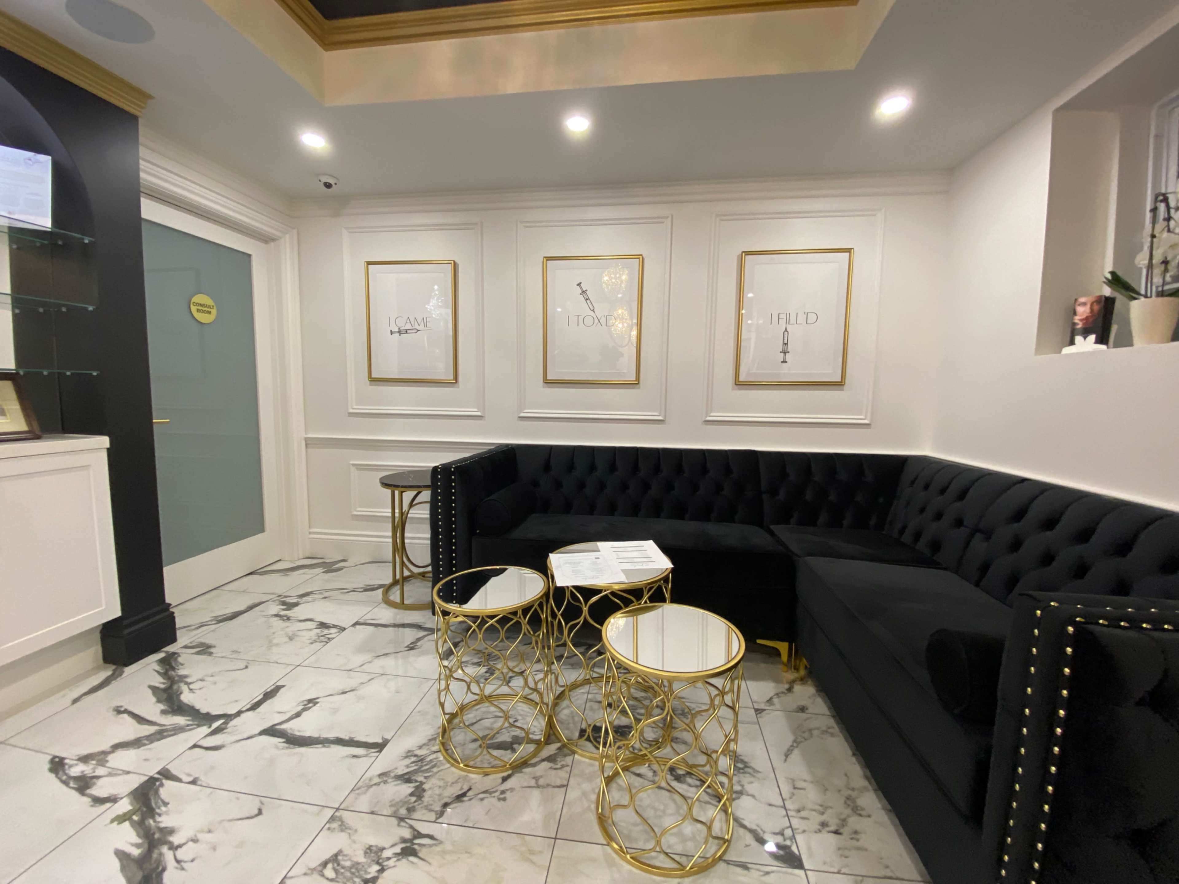 A modern waiting area with a black velvet sofa, marble flooring, and gold-accented tables, along with framed artwork on the walls.