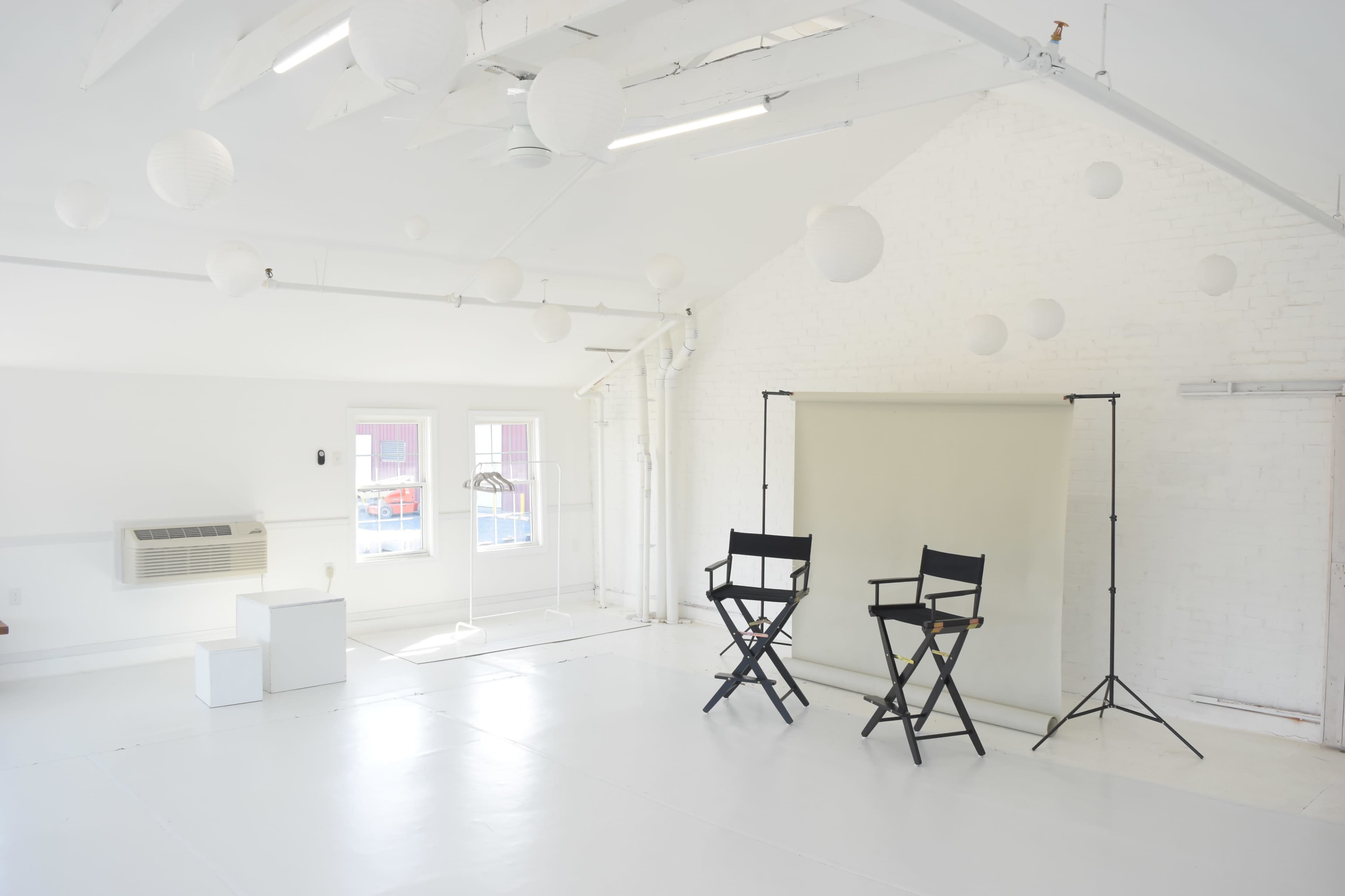 The image shows a bright, white studio space featuring two director's chairs, a backdrop on a stand, and decorative paper lanterns hanging from the ceiling.