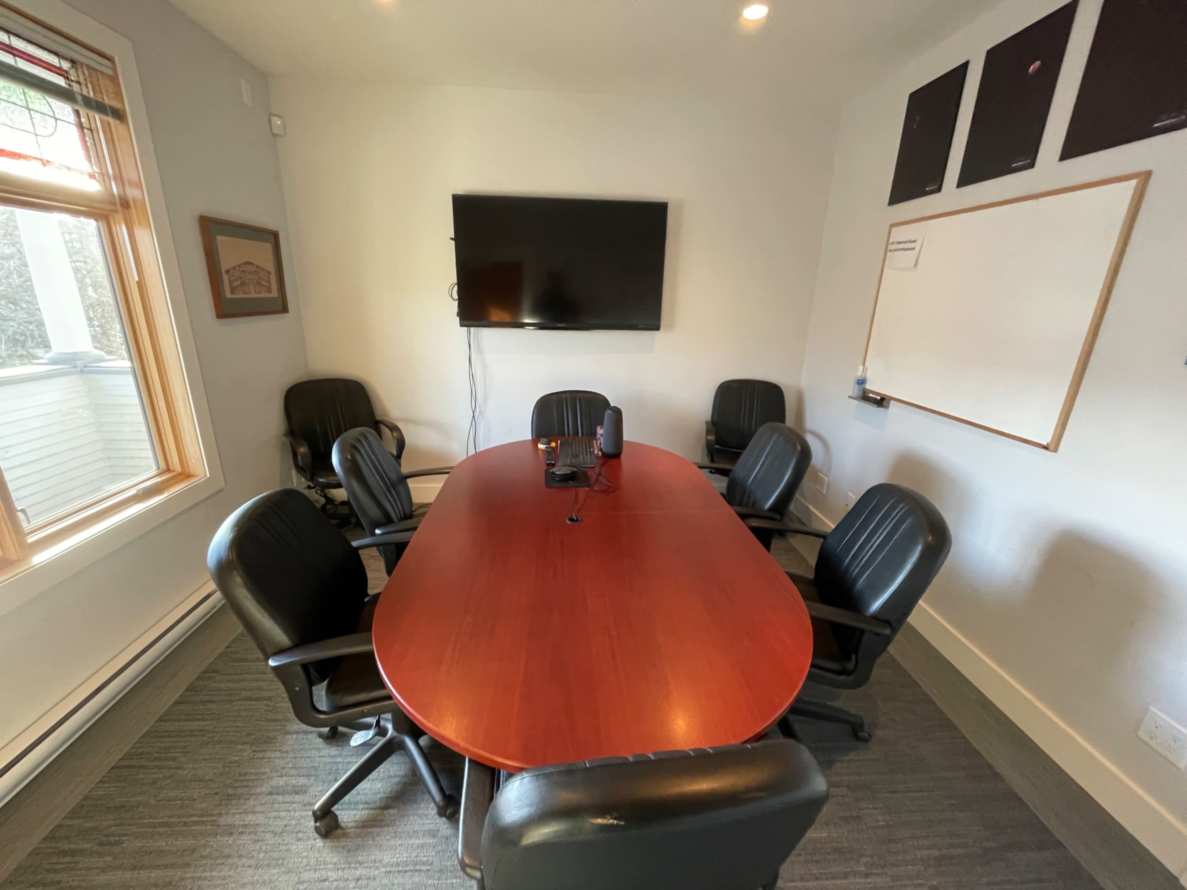 A conference room features a large oval table surrounded by six black chairs, with a wall-mounted television and a whiteboard.