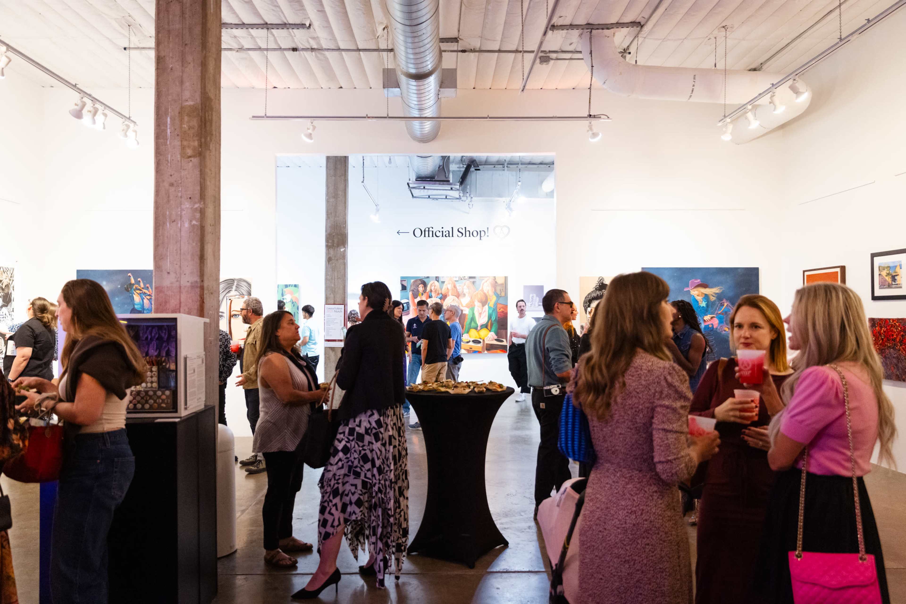 A crowded art gallery features people chatting and browsing artworks while a sign for an official shop is displayed in the background.