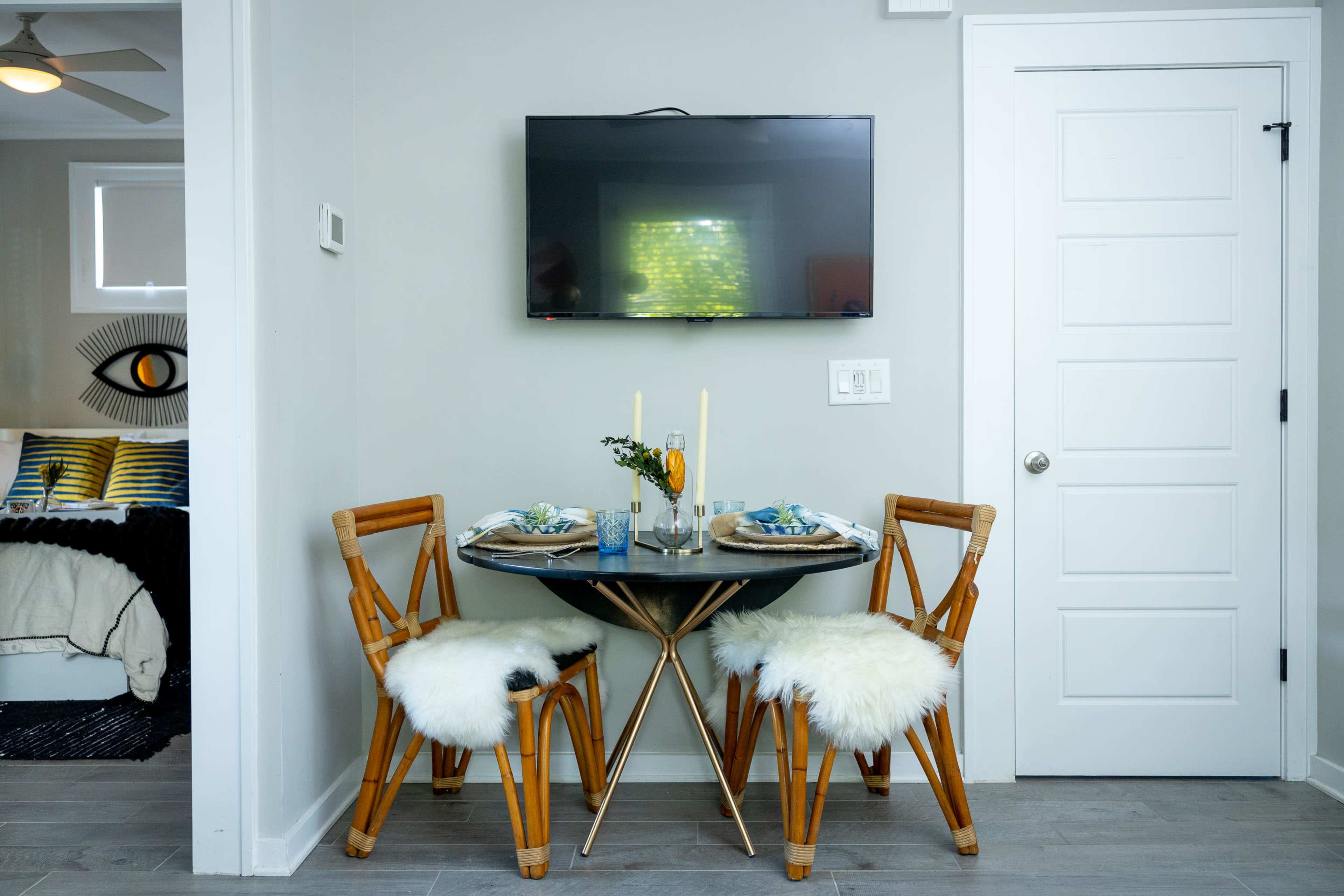 A small dining area features a round black table with two wooden chairs adorned with white fur throws, set for a meal under a wall-mounted television.