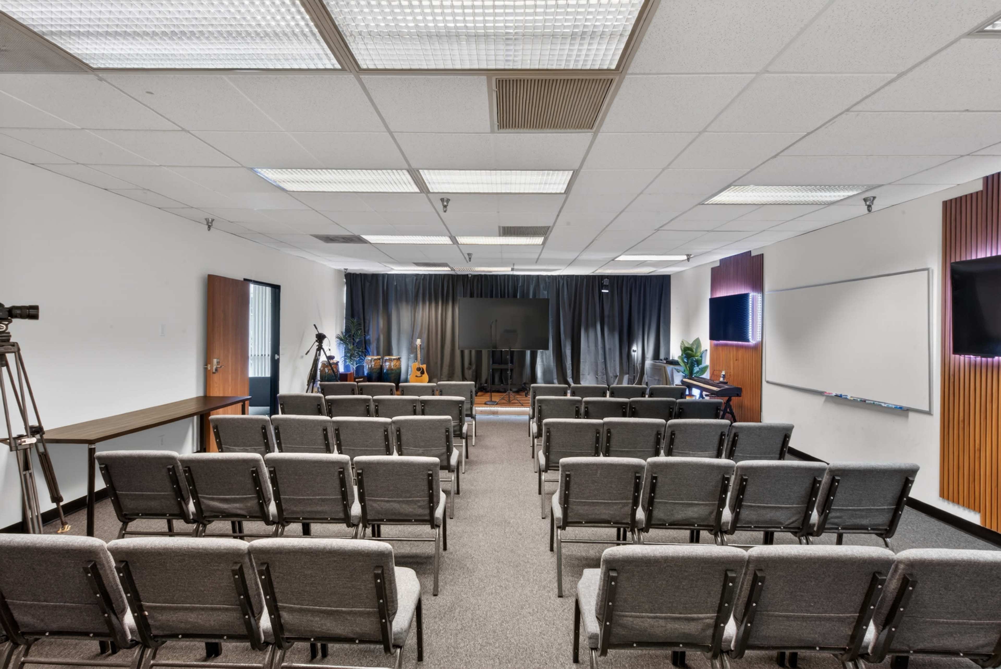 The image shows a conference room arranged with rows of grey chairs facing a stage with a dark backdrop, flanked by cameras and screens.