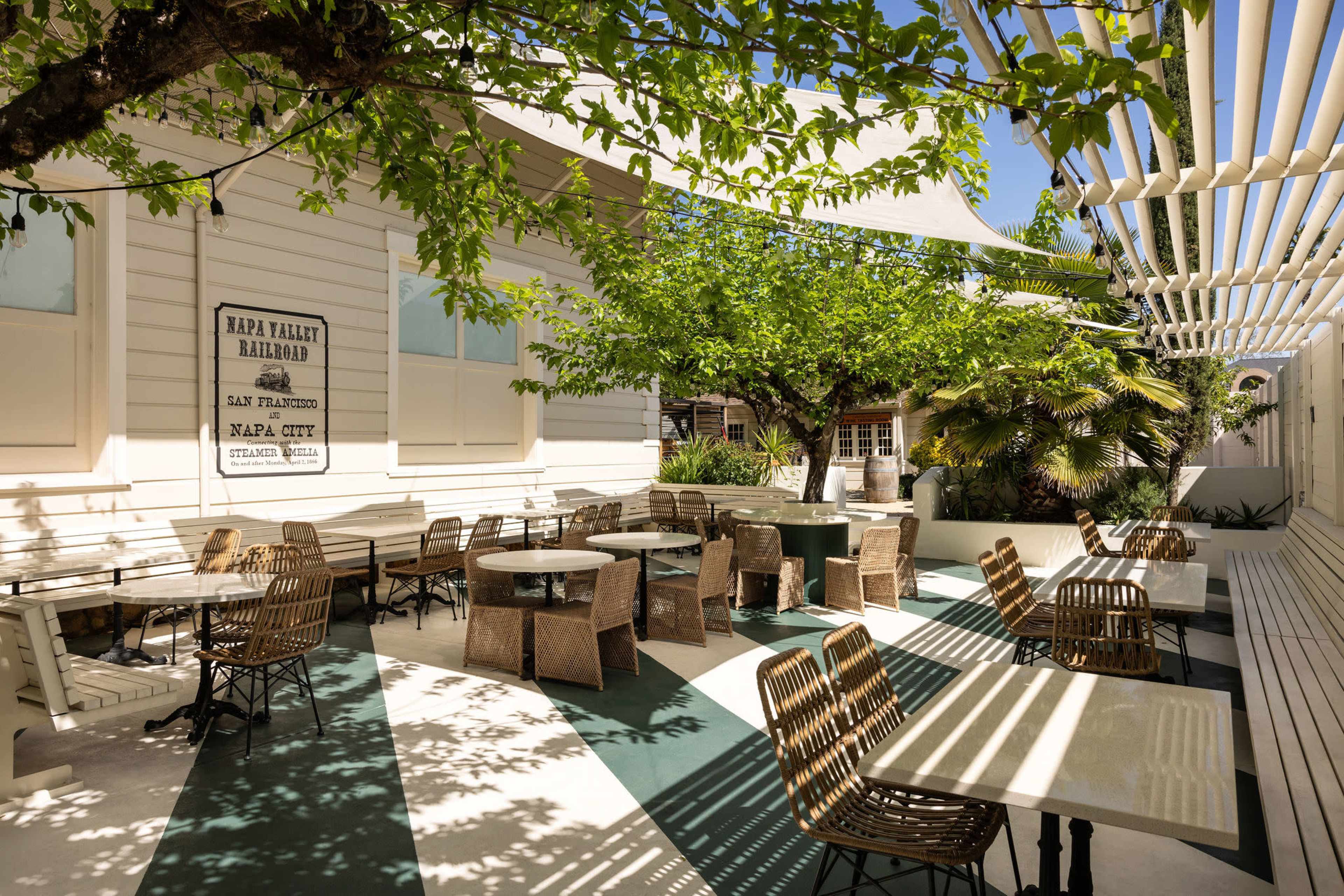 The image shows a patio area with several tables and chairs surrounded by greenery and shade structures.
