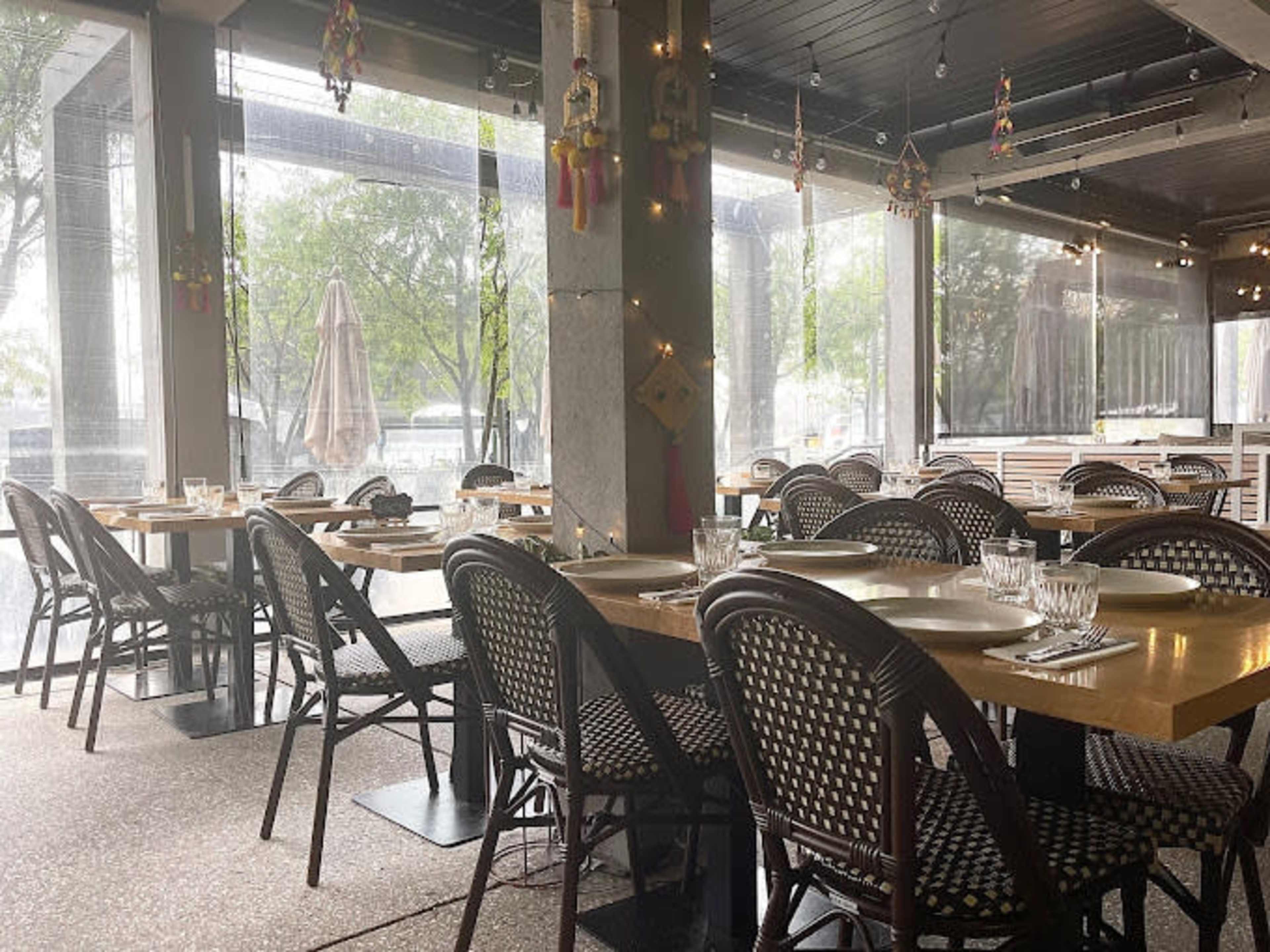 The image shows an empty restaurant with wooden tables and woven chairs, illuminated by natural light filtering through large windows.
