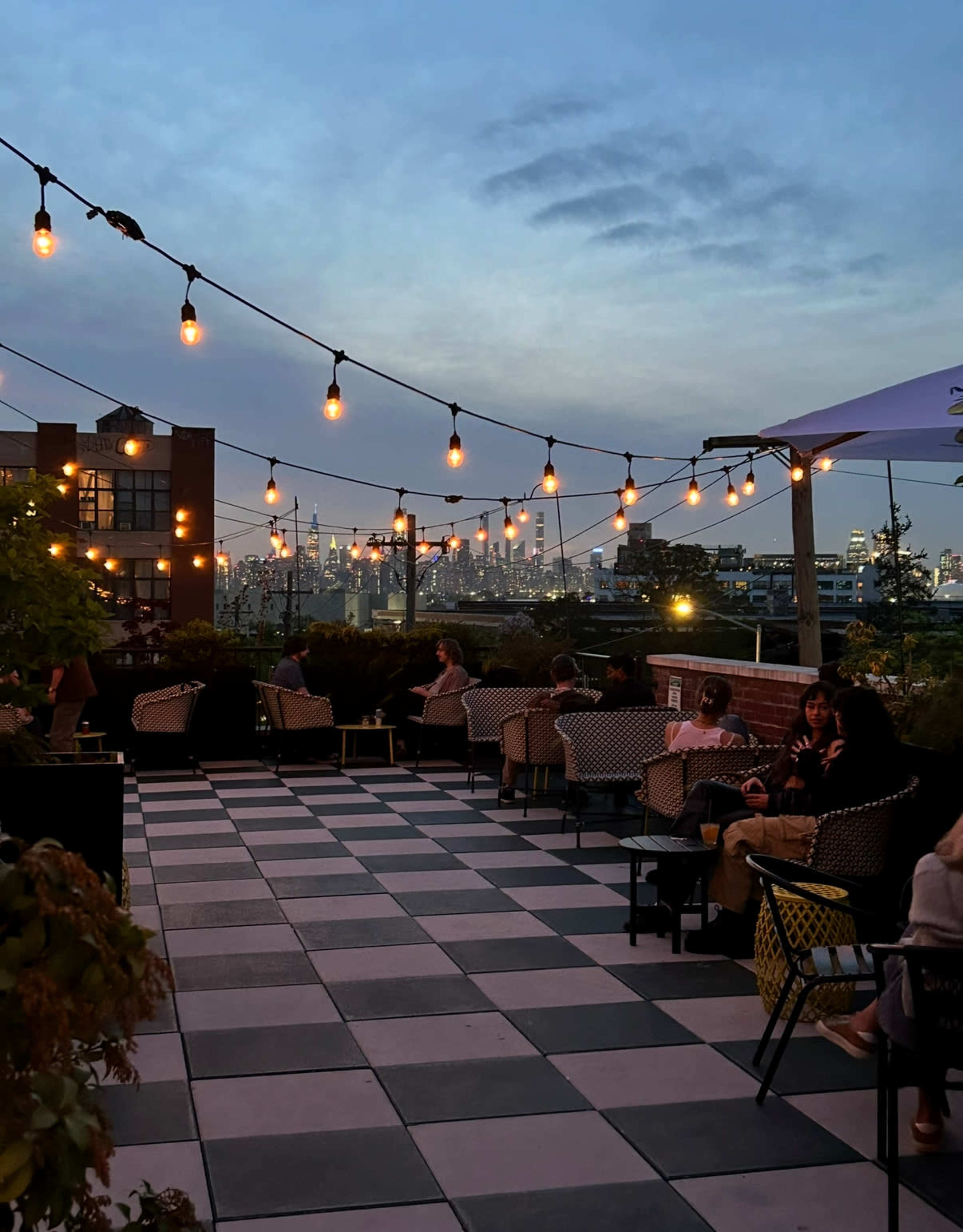 A rooftop bar with string lights overhead and patrons seated on a checkered floor, overlooking a city skyline at dusk.