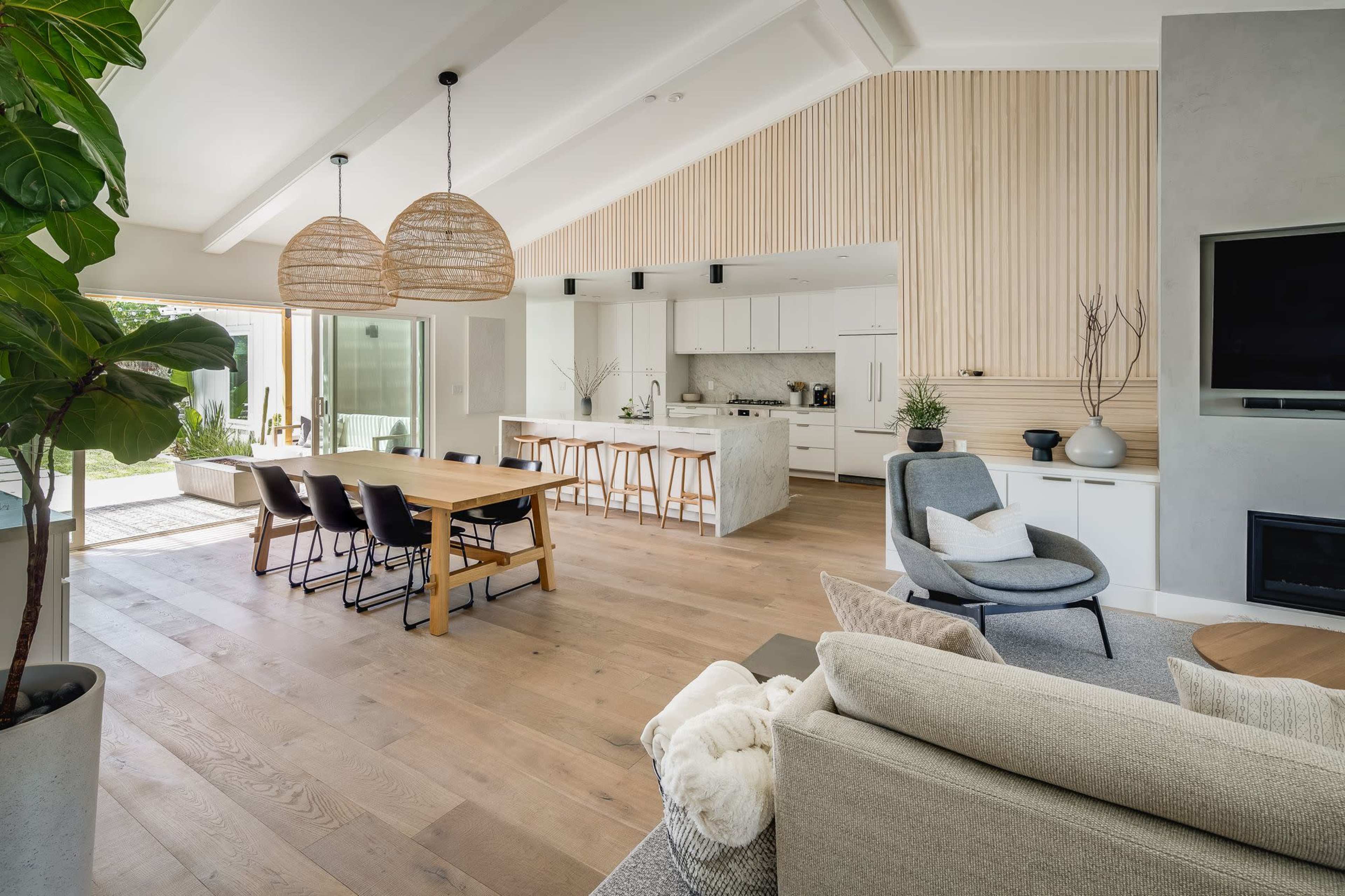 A spacious, modern open-concept living and dining area with a wooden dining table, comfortable seating, and a well-lit kitchen in the background.