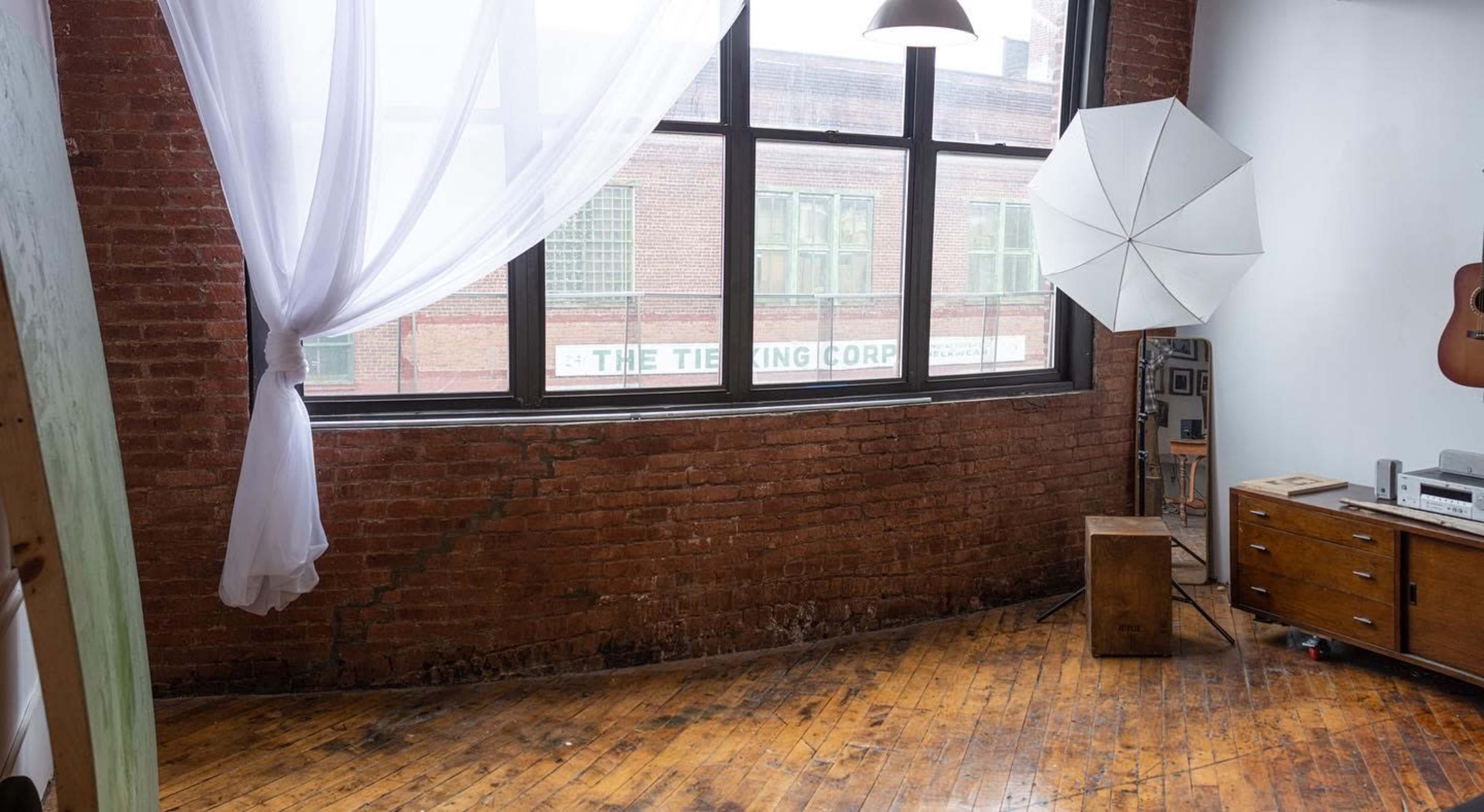 A corner of a room with exposed brick walls, a large window draped with sheer fabric, a light umbrella, and wooden flooring.