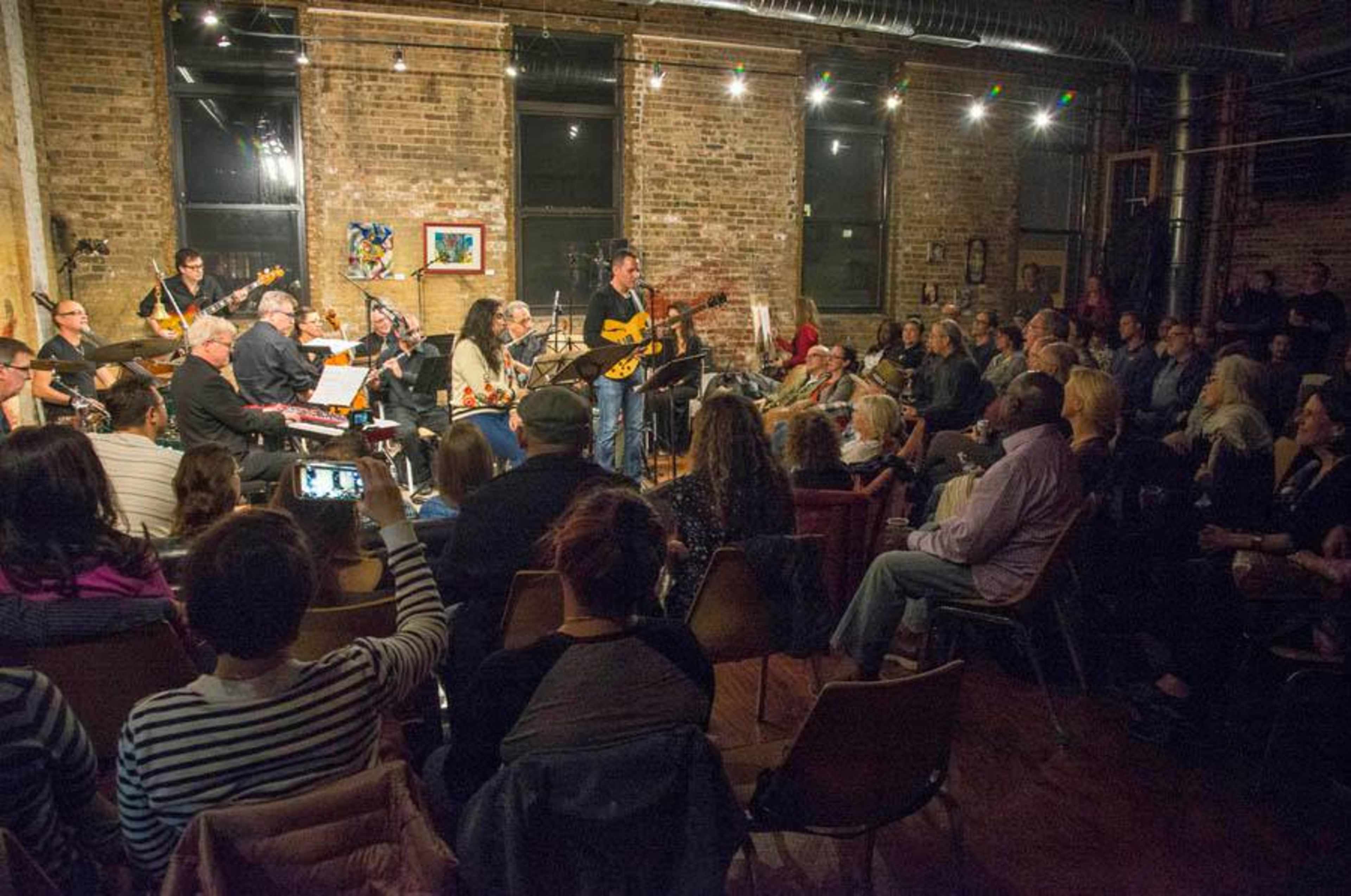 A musician performs on stage in front of a seated audience in a brick-walled venue with various instruments and a band behind him.