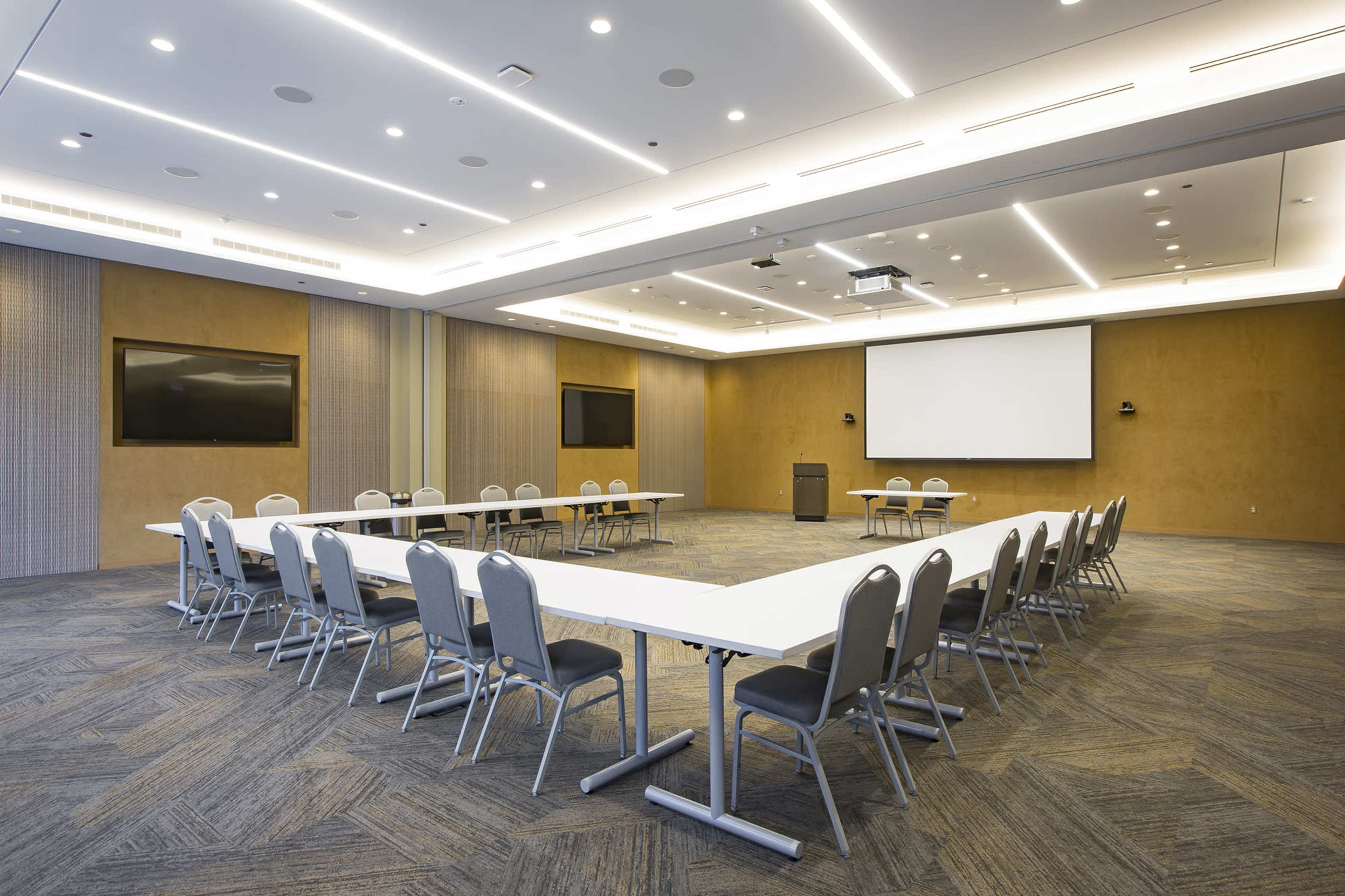 A spacious conference room features a large, U-shaped arrangement of tables and chairs, with a projector screen and two monitors on the walls.