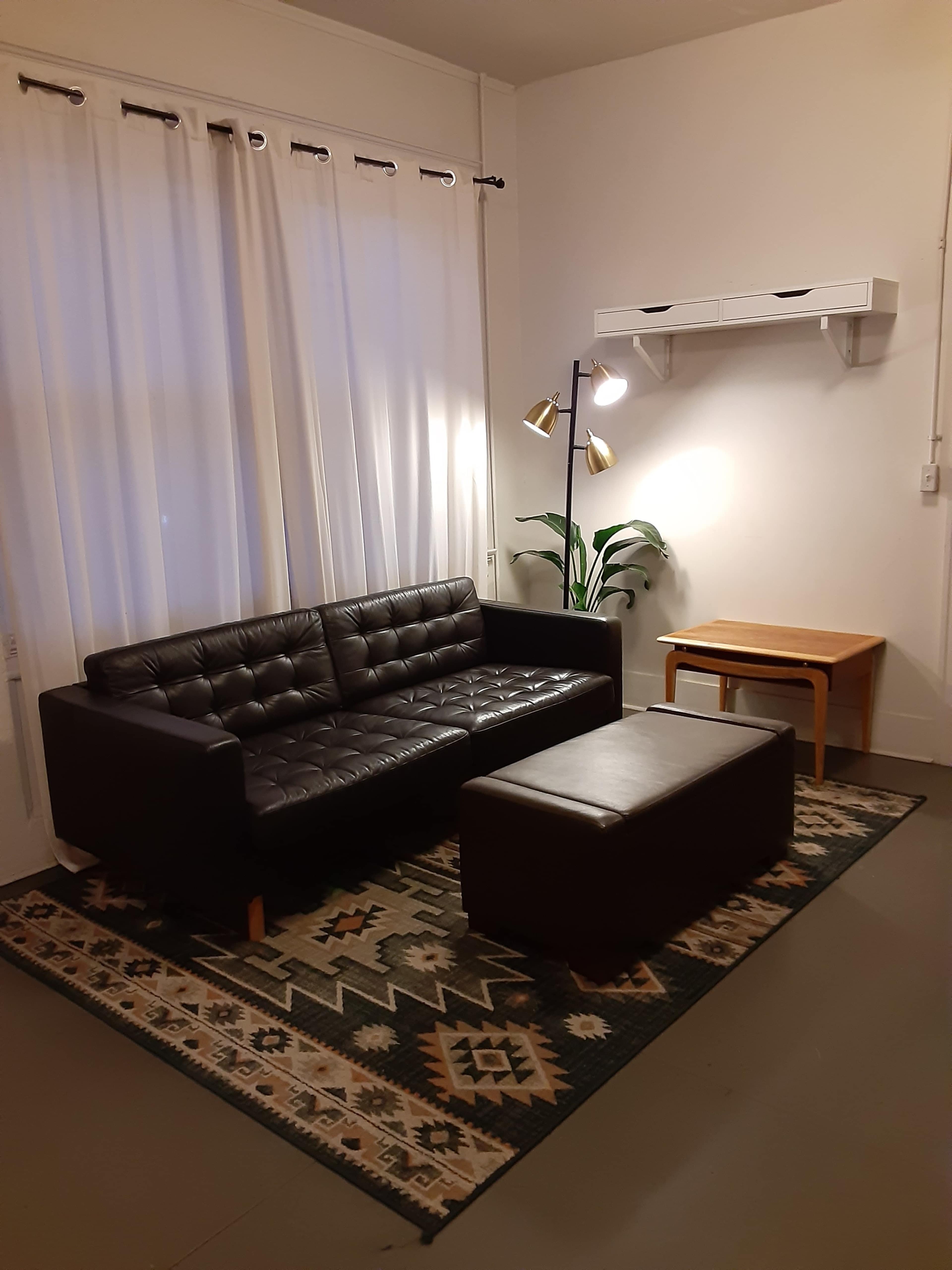 A simple living room featuring a black leather sofa, an ottoman, a small table, and a floor lamp beside a potted plant, all arranged on a patterned rug under large, white curtains.