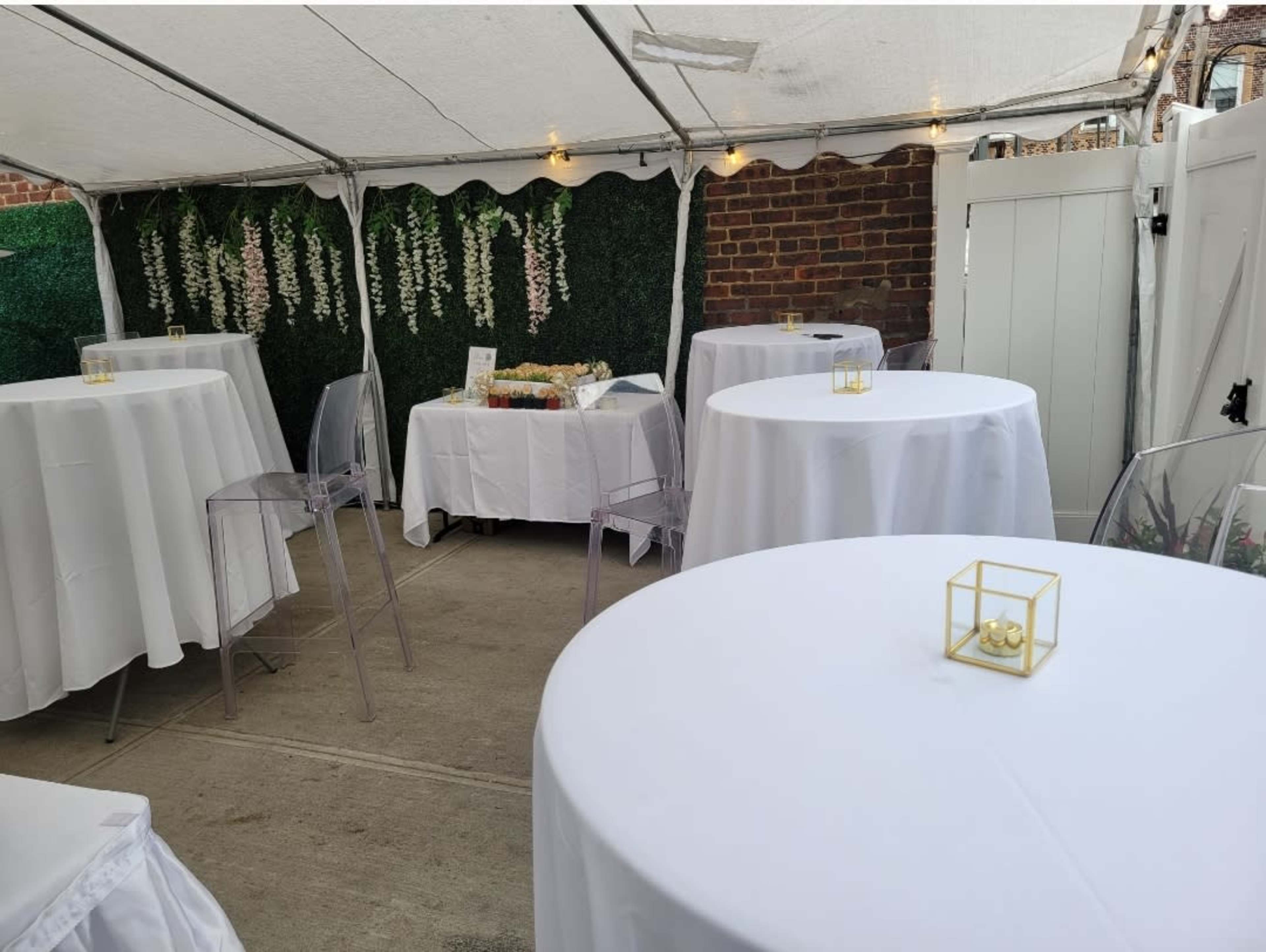 A tented area features several round tables with white tablecloths and transparent chairs, set against a backdrop of greenery and brick walls.