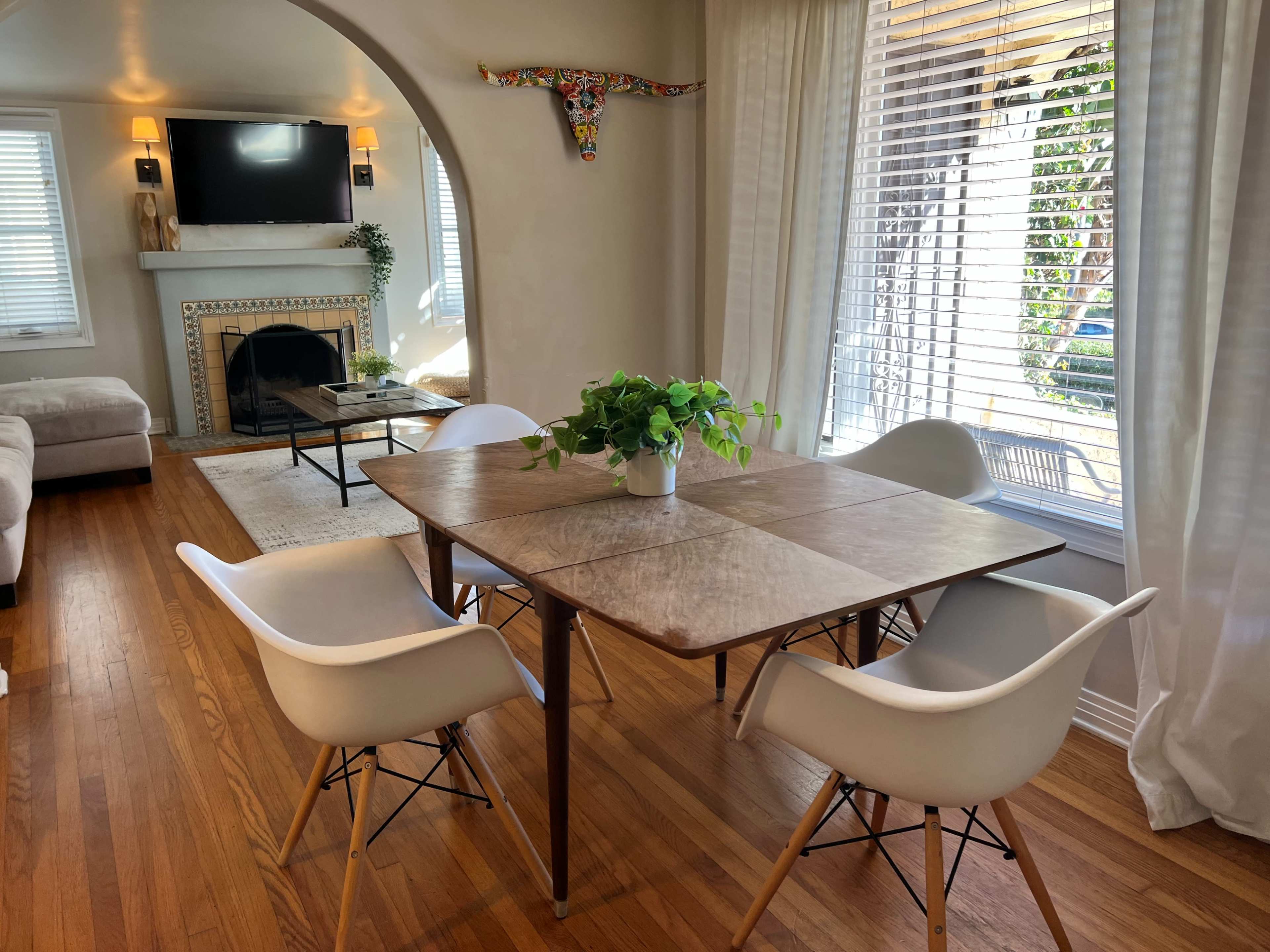 A dining area features a square wooden table surrounded by four white chairs, with a decorative plant in the center, and a living room visible in the background.