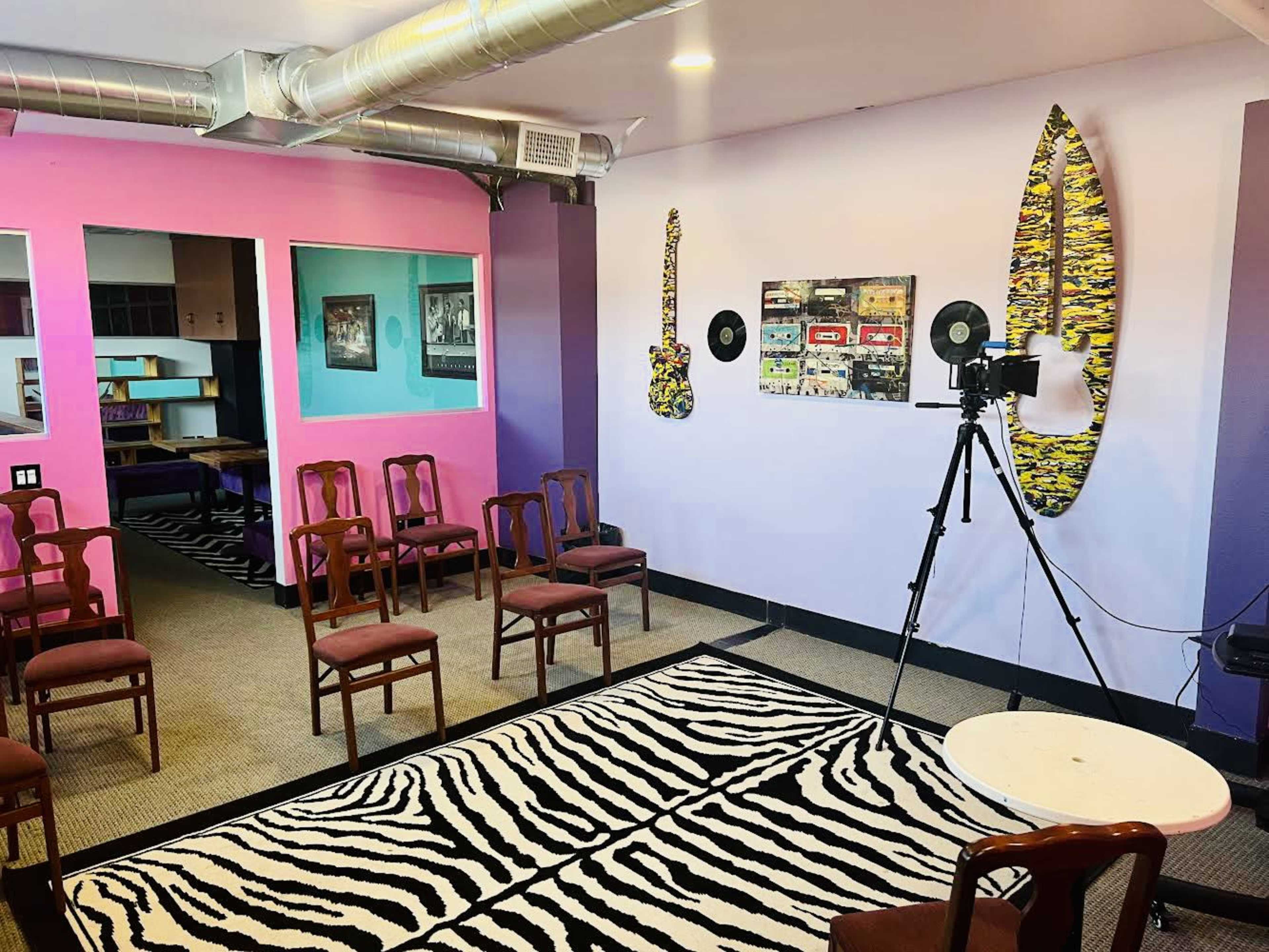 A vibrant room features several chairs arranged in a semi-circle, a camera on a tripod, and decorated walls with colorful artwork and surfboards.