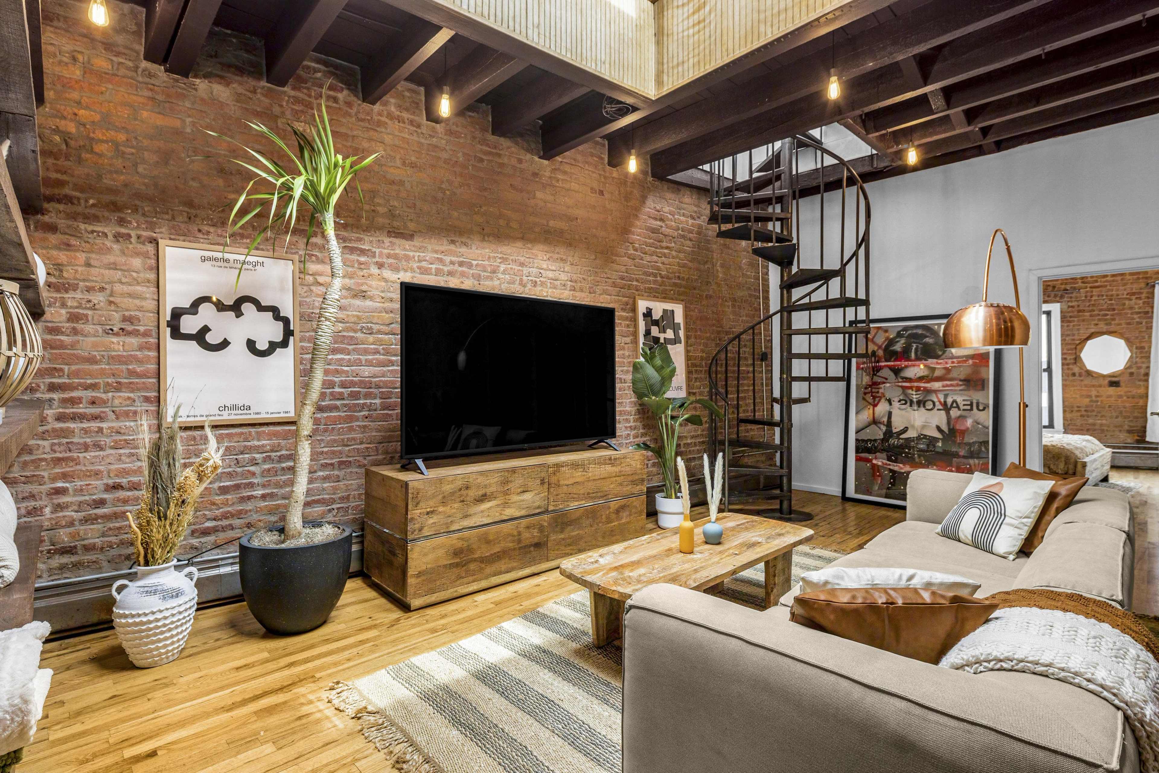 A stylish living room featuring exposed brick walls, a comfortable sofa, a large TV on a wooden stand, and a spiral staircase leading to an upper level.