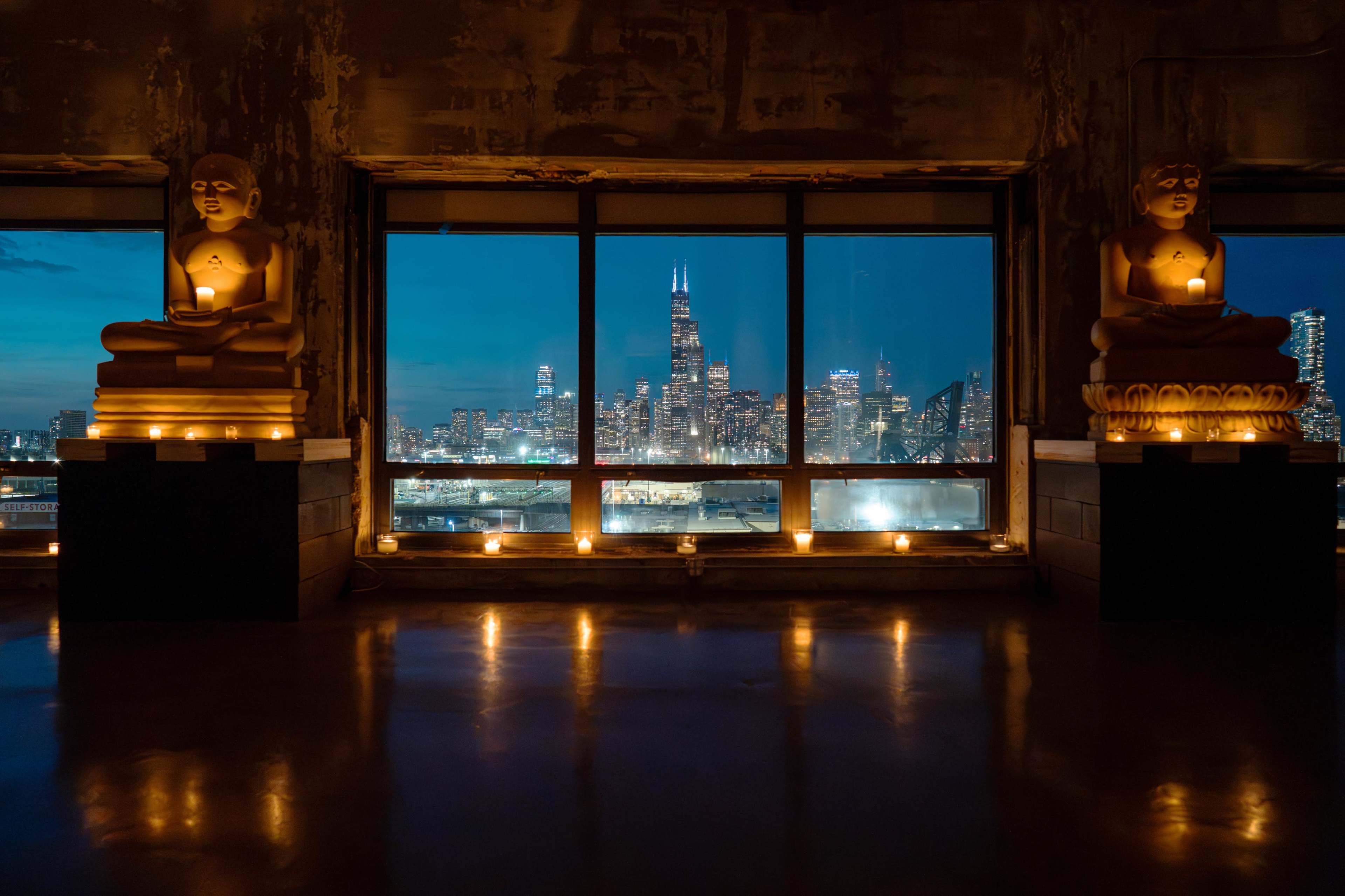 The scene shows a dimly lit room with two Buddha statues on pedestals, candles placed around them, and a panoramic view of the Chicago skyline through large windows at dusk.
