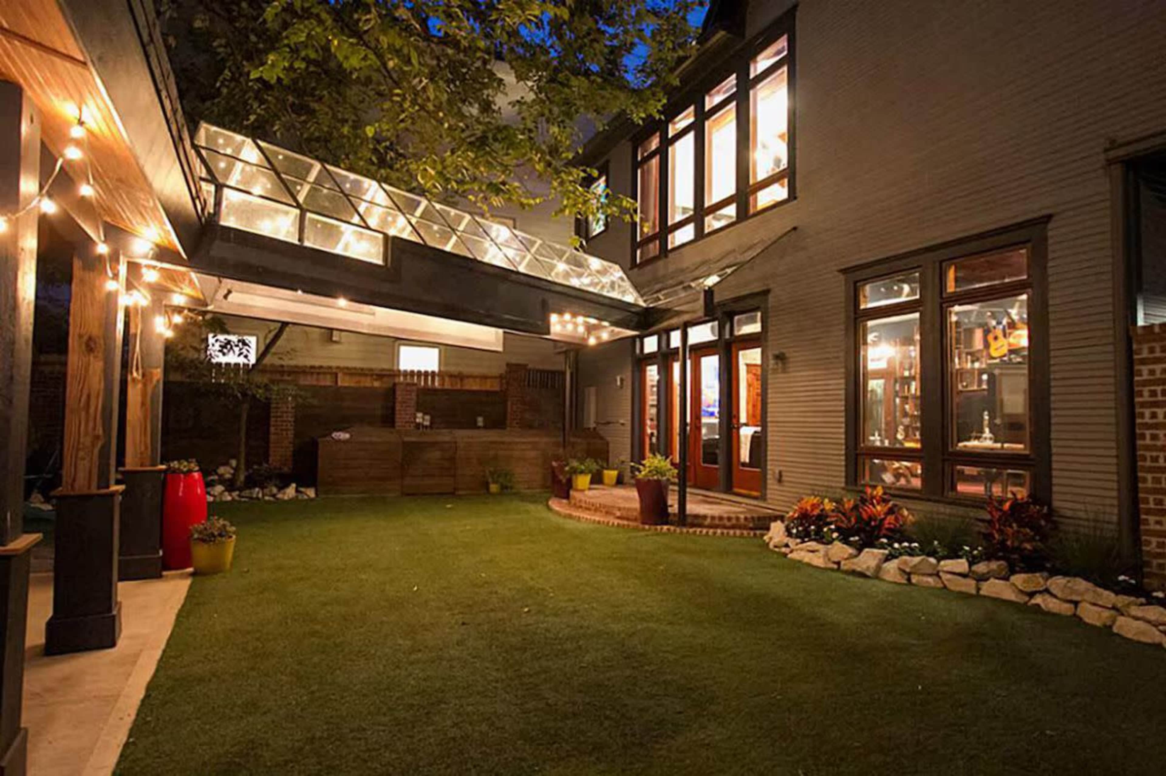 A well-lit backyard features a modern patio with overhead glass panels and neatly arranged garden beds.