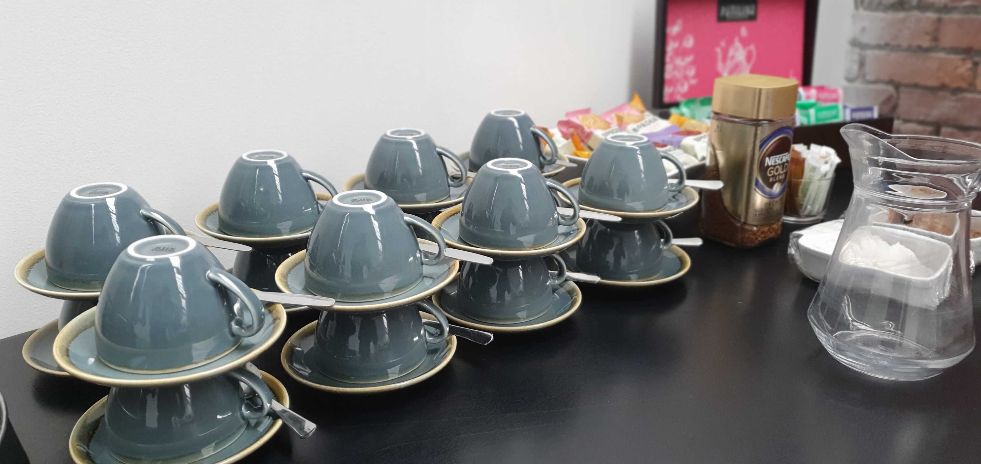 The image shows a row of stacked coffee cups on a table, accompanied by a variety of beverage condiments and packaging in the background.