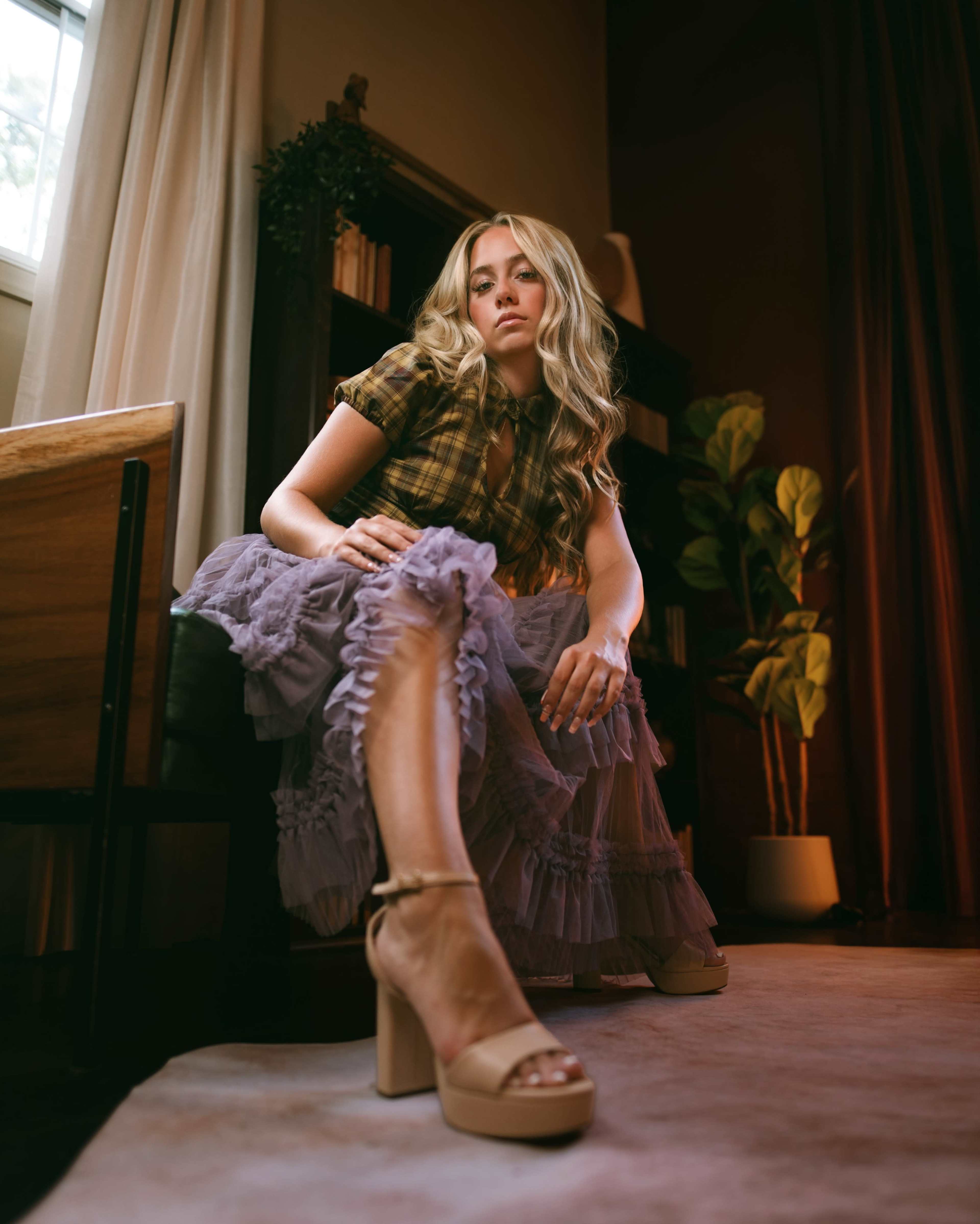 A person sits on a chair wearing a plaid top and a layered purple skirt, with a potted plant visible in the background.