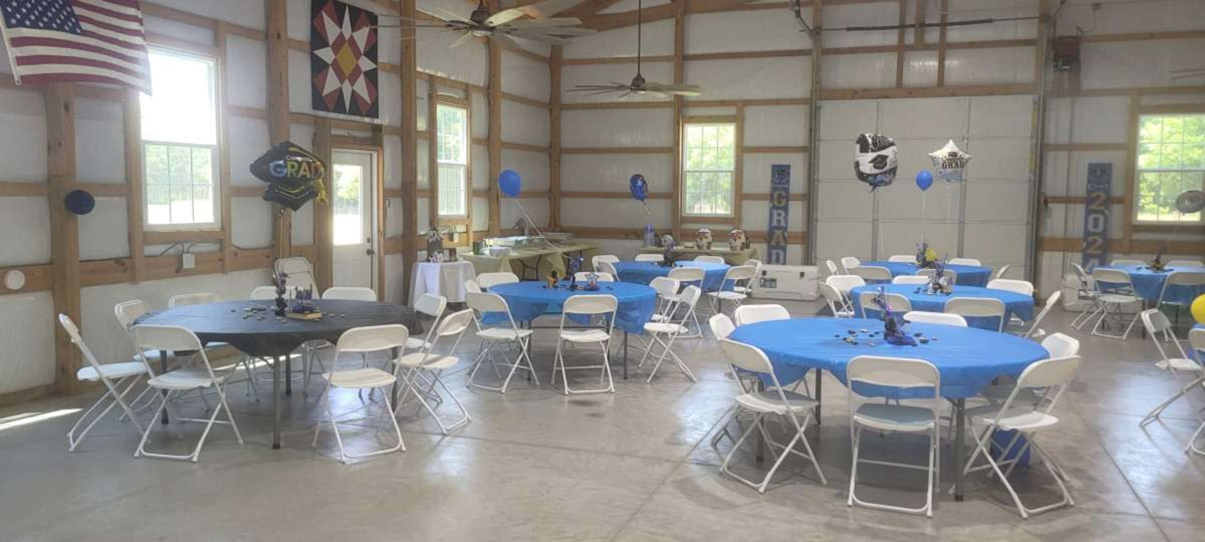 The image shows a spacious indoor venue with several tables set for a celebration, featuring blue tablecloths and decorations, and an American flag hanging on one wall.