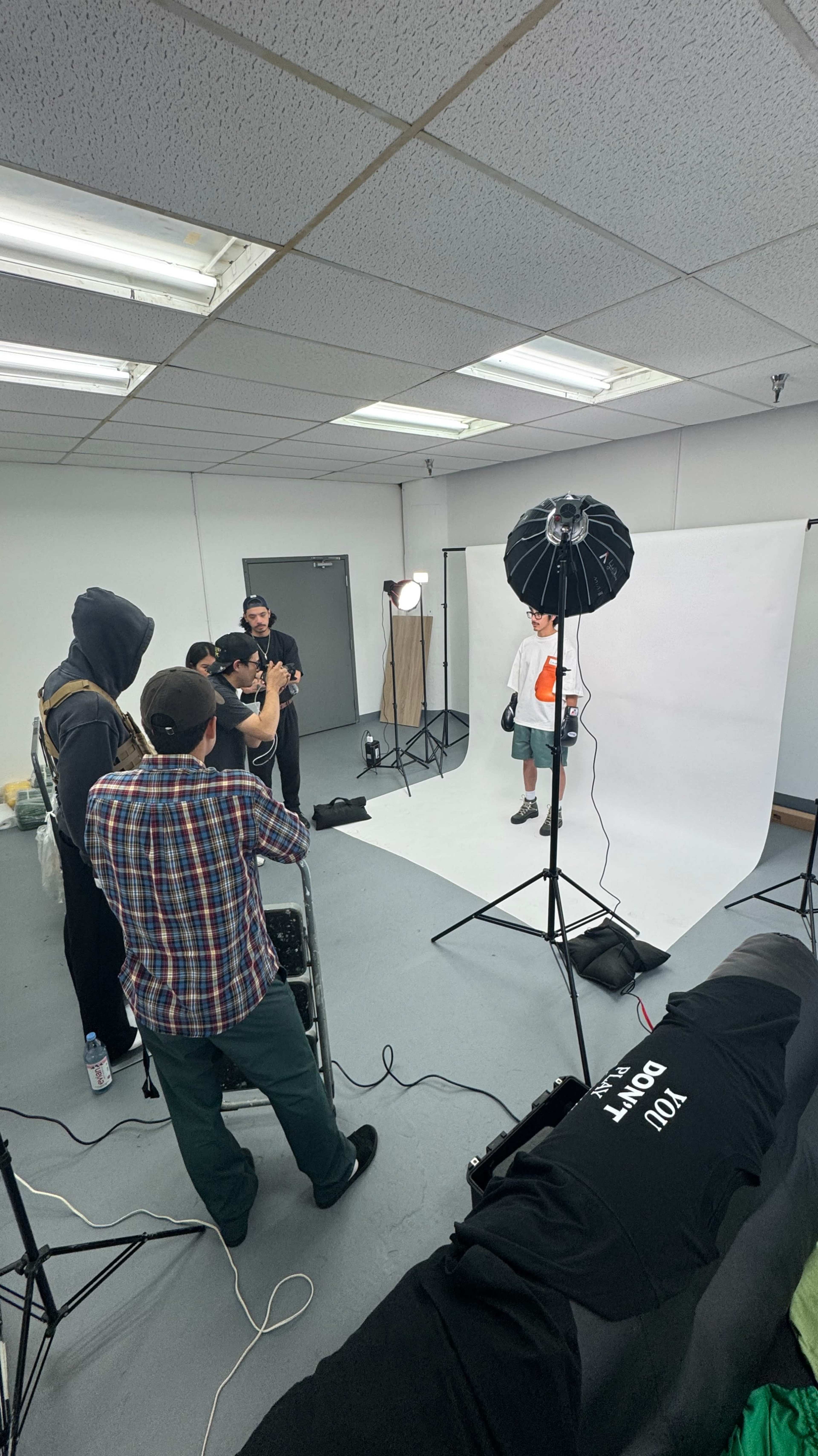 A group of five individuals is gathered in a studio, preparing for a photoshoot with a model in front of a white backdrop and lighting equipment.