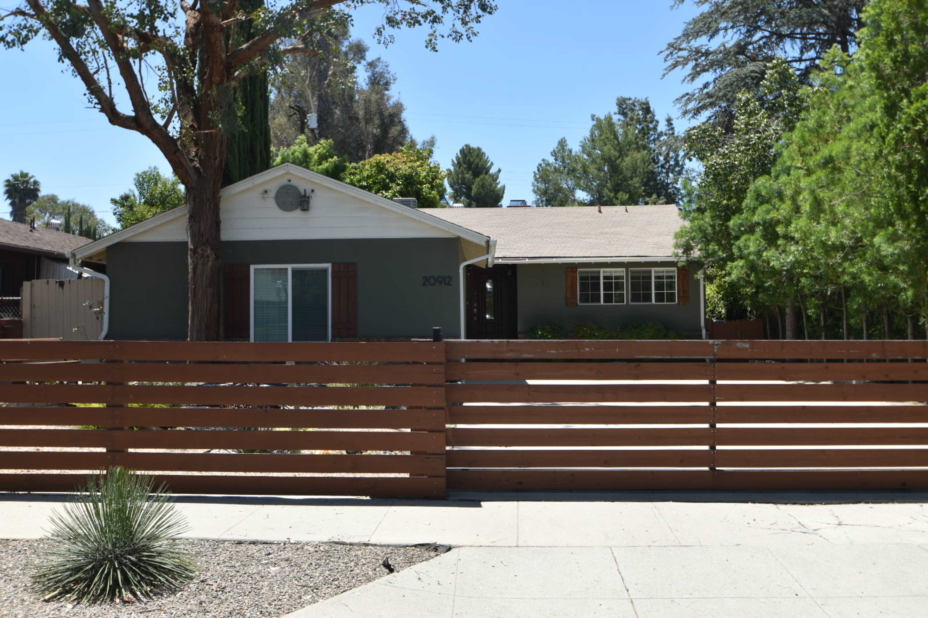 A single-story gray house with a fenced front yard sits under a clear blue sky.