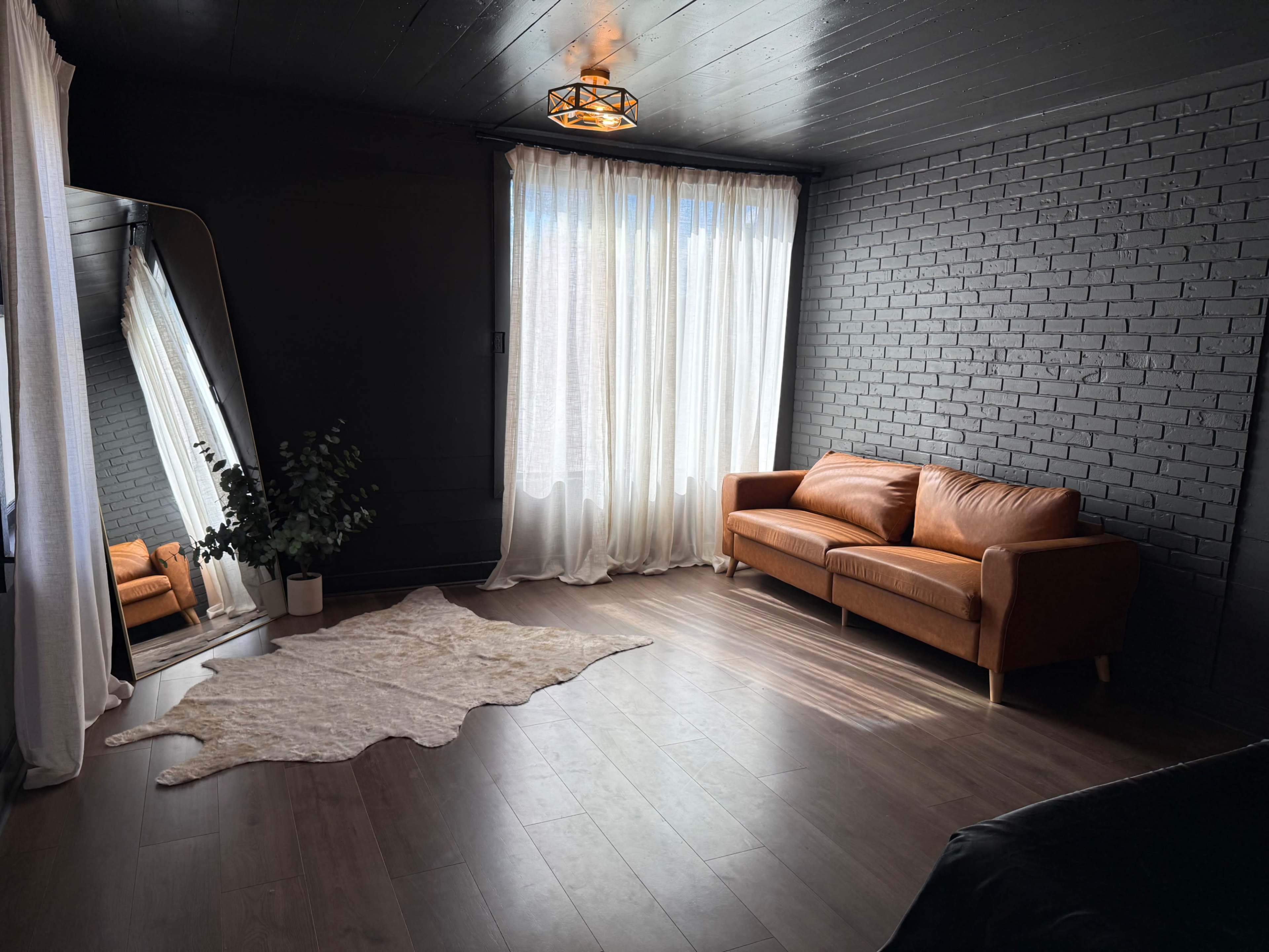 The image shows a minimalist interior with a leather sofa, a large mirror, sheer curtains, and a potted plant, all set against dark brick walls and wooden flooring.