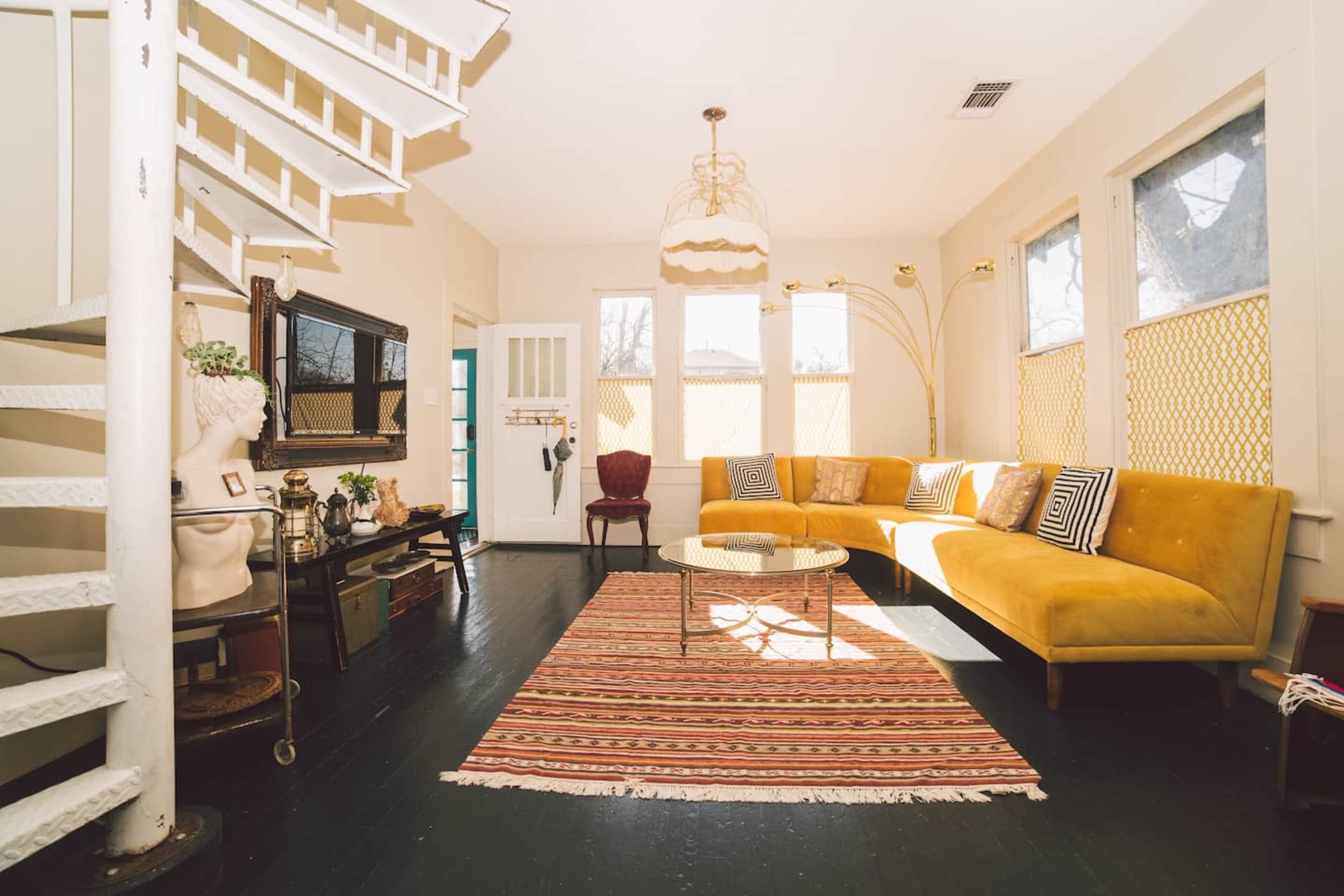 A bright living room features a yellow sectional sofa, a round glass coffee table on a patterned rug, and a staircase with a white railing.