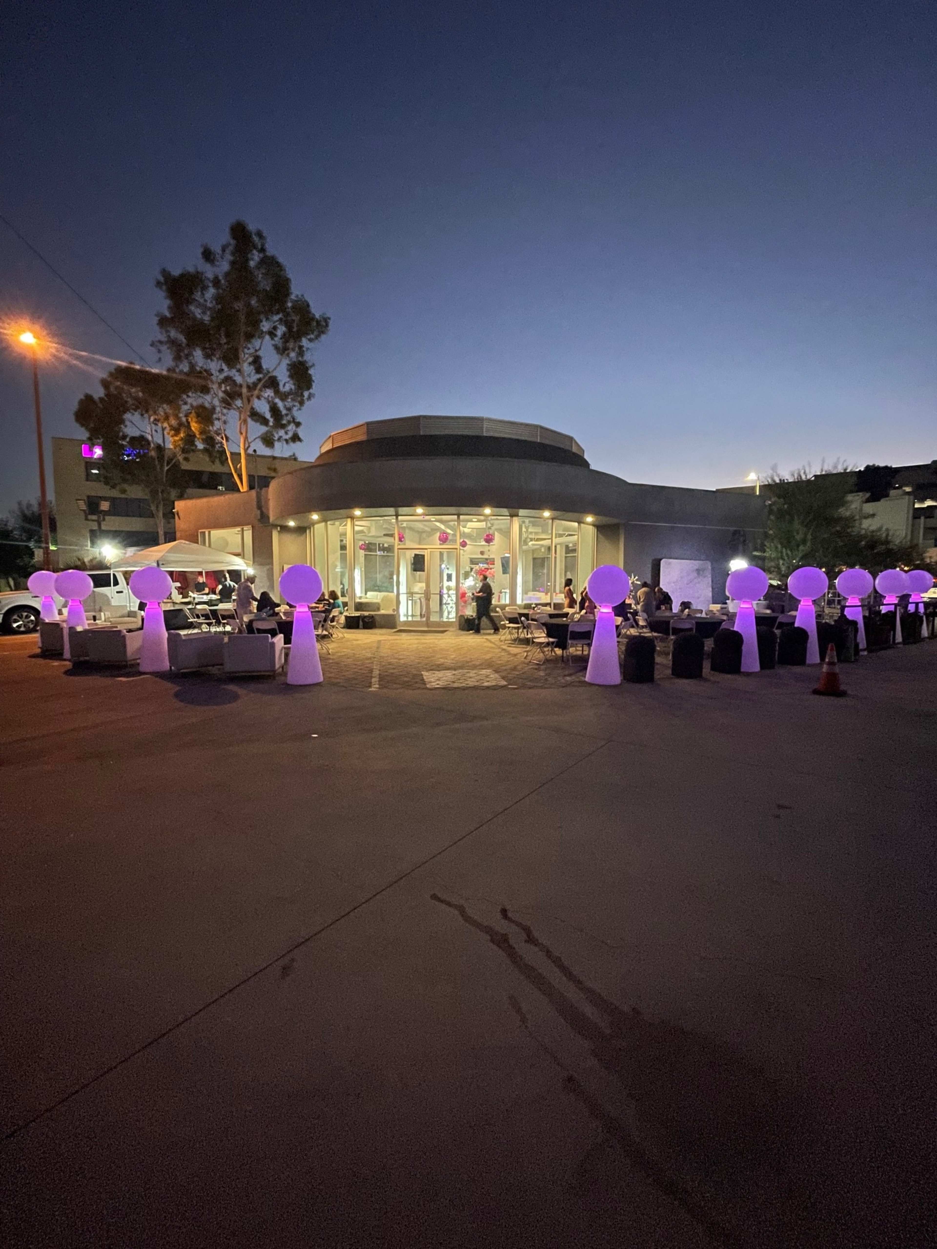 A modern building with large glass windows is illuminated by purple lights, surrounded by lighted sculptures and people seated outside in the evening.