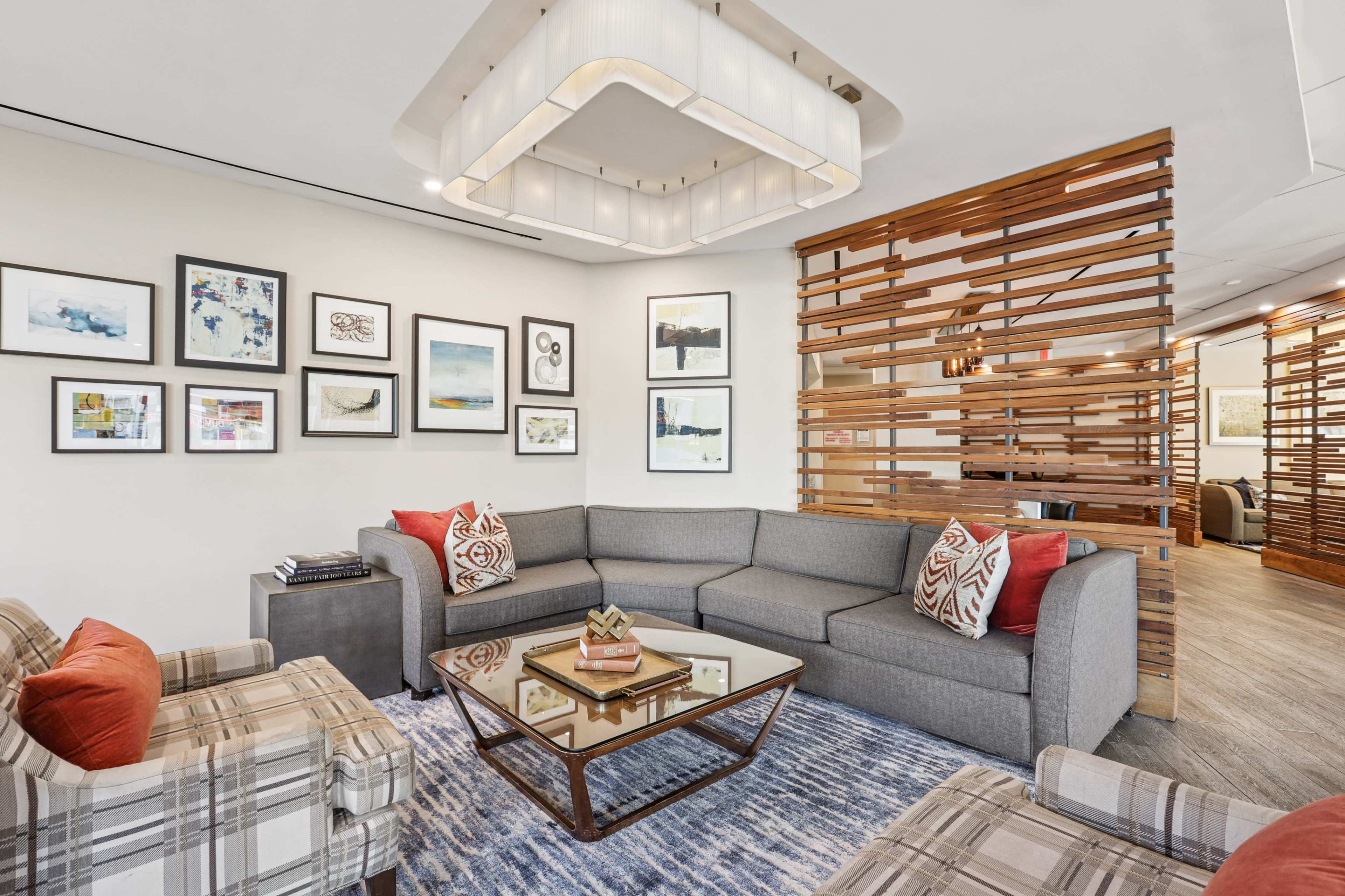 The image shows a modern living room featuring a gray sectional sofa, a glass coffee table, and a wooden partition with framed artwork on the walls.