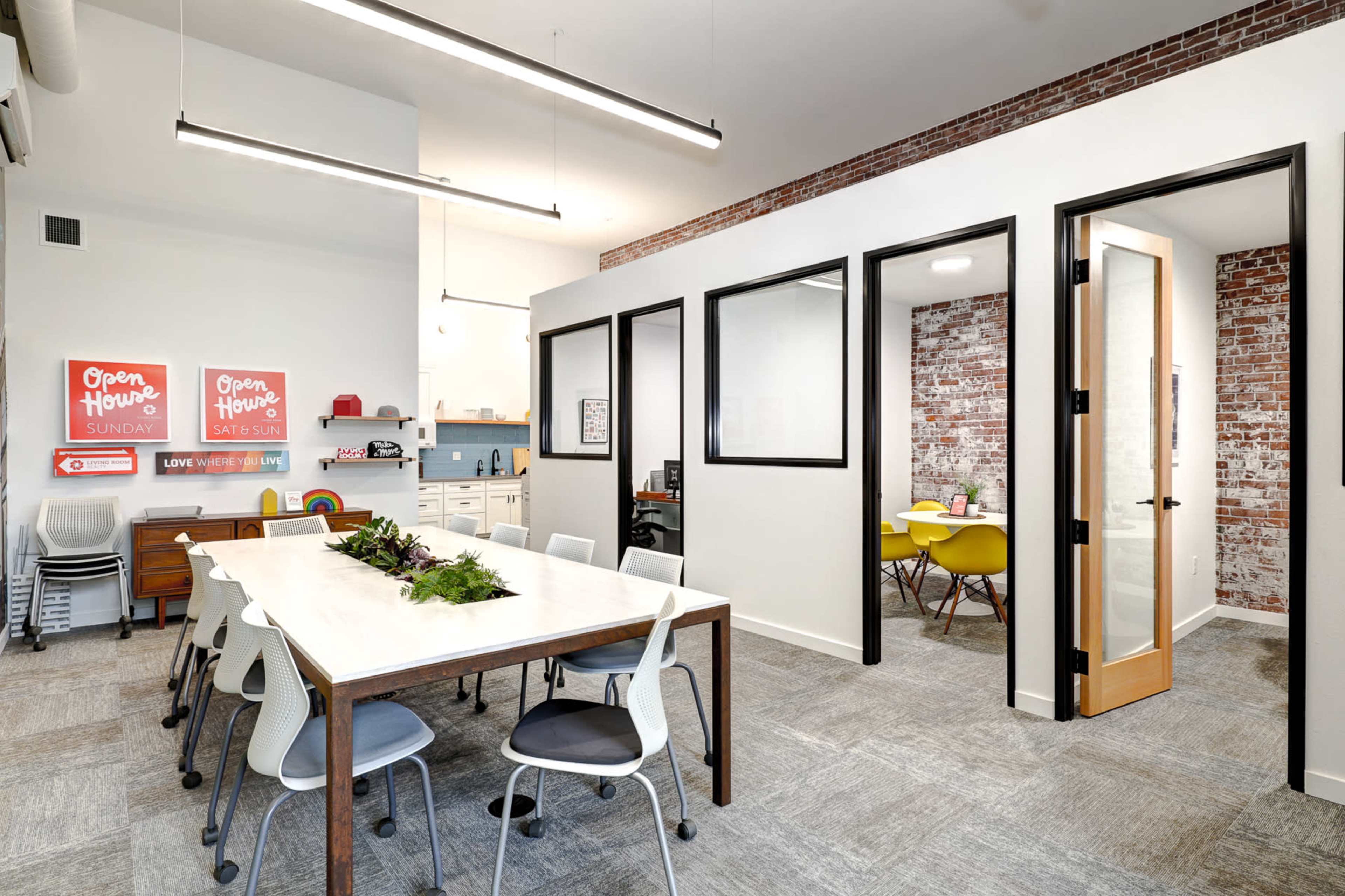 A bright office space features a large table surrounded by chairs, with a kitchenette area and glass-partitioned meeting rooms in the background.