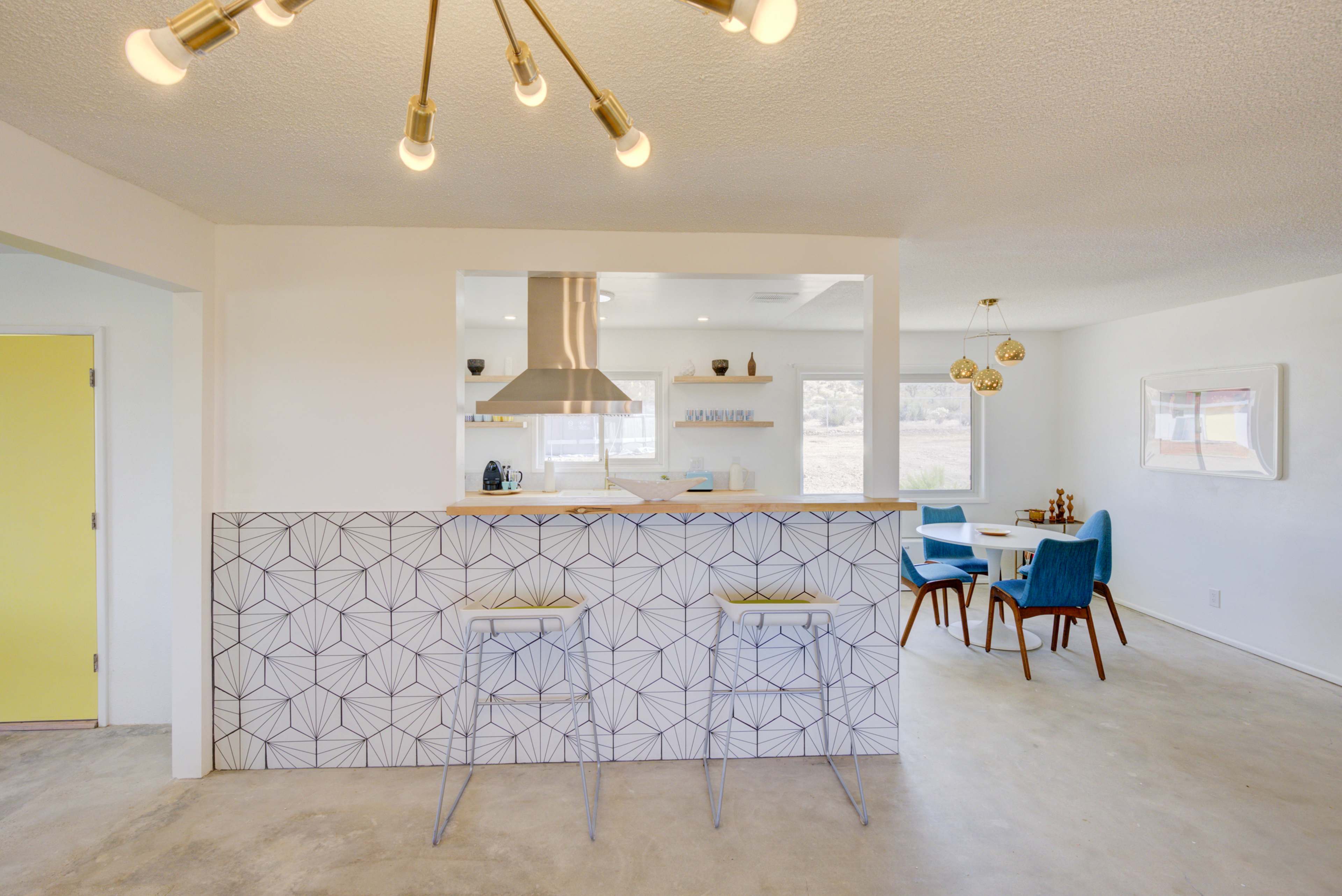 A modern kitchen with a geometric patterned bar separates the cooking area from a dining space, featuring a round table and colorful chairs.