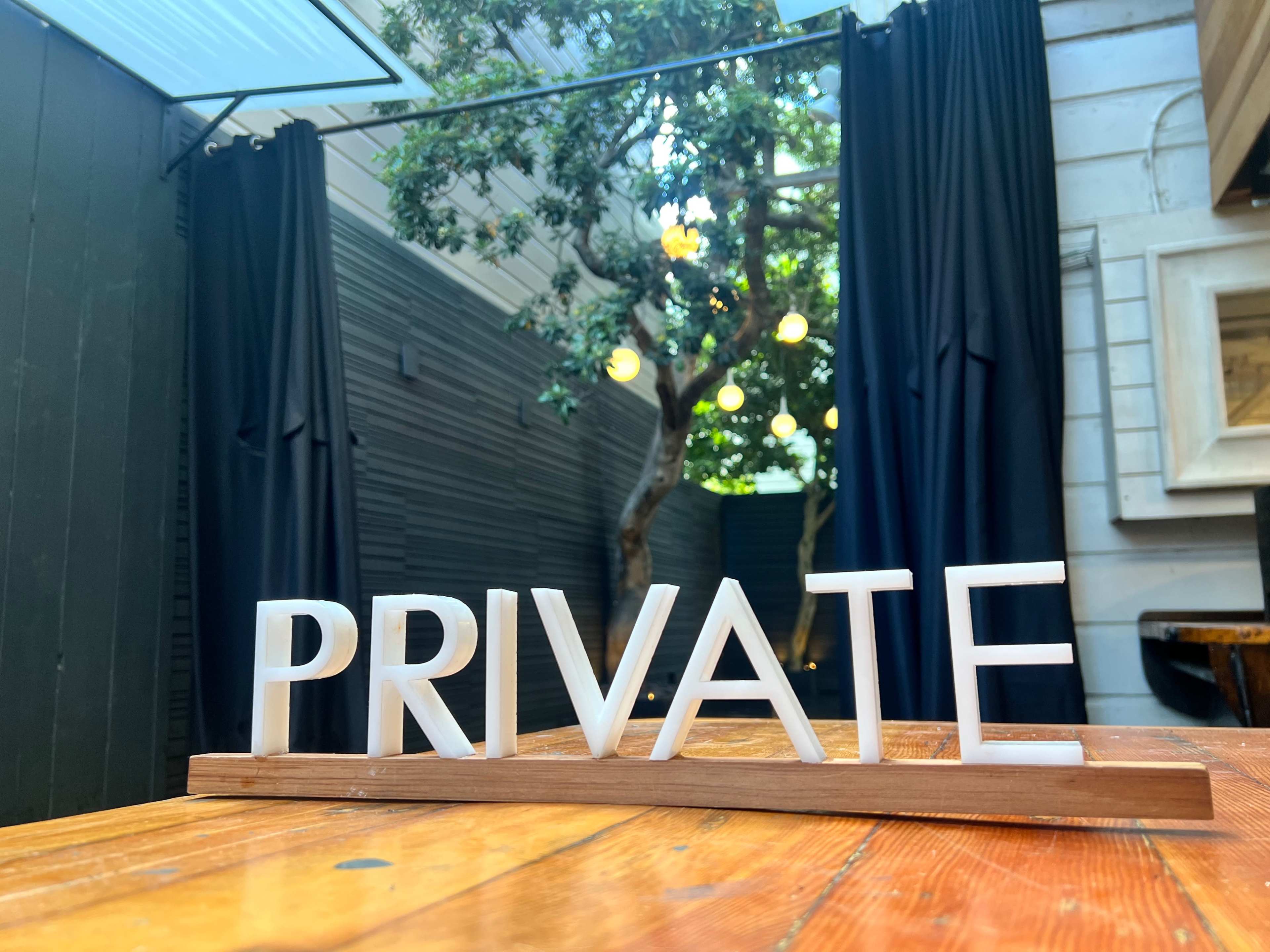 A wooden sign spelling "PRIVATE" placed on a table, with a backdrop of dark curtains and a tree adorned with lights.